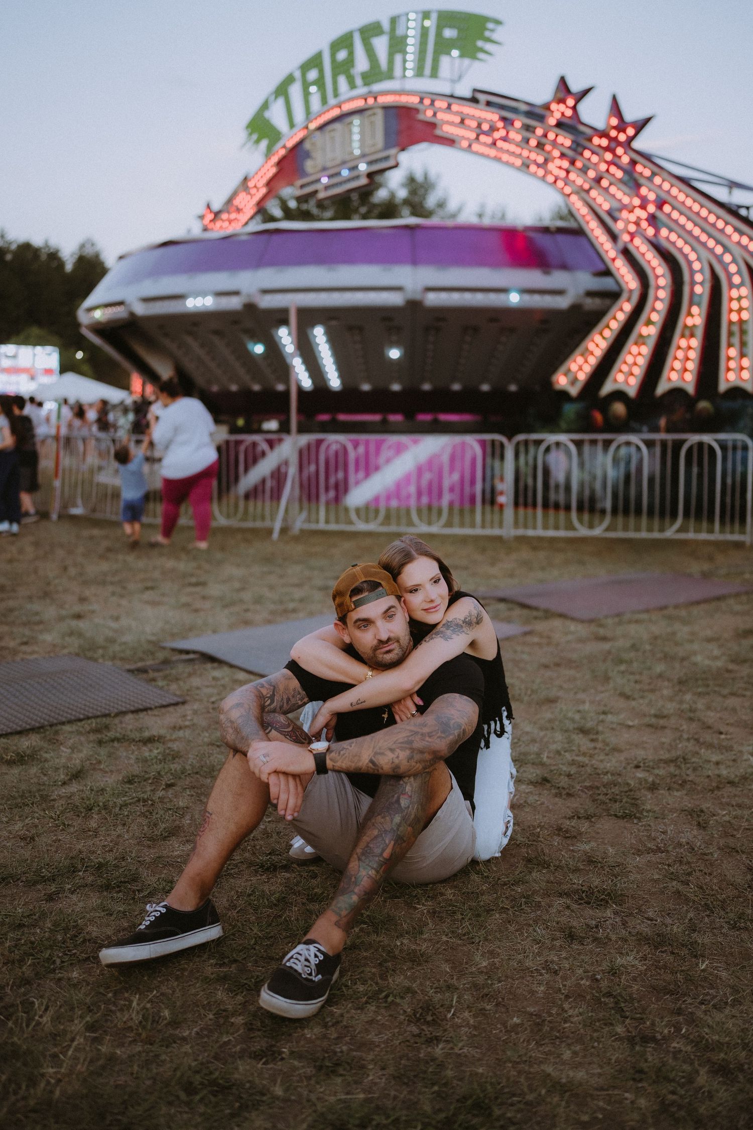 Couple sitting together on grass at a fairground, with colorful Starship ride lights glowing behind them at dusk.