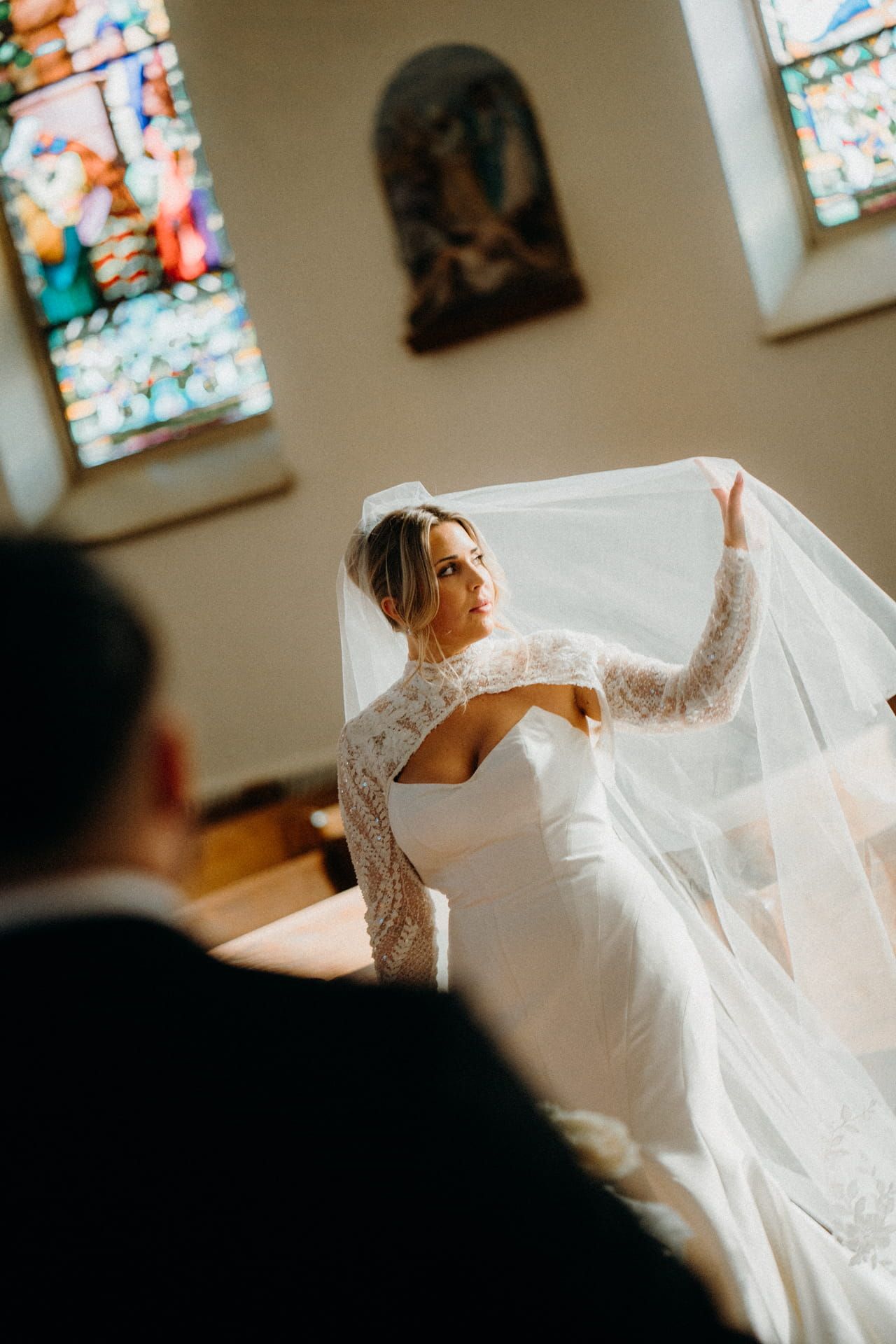 Bride in lace wedding gown holding veil in sunlit church with stained glass windows.