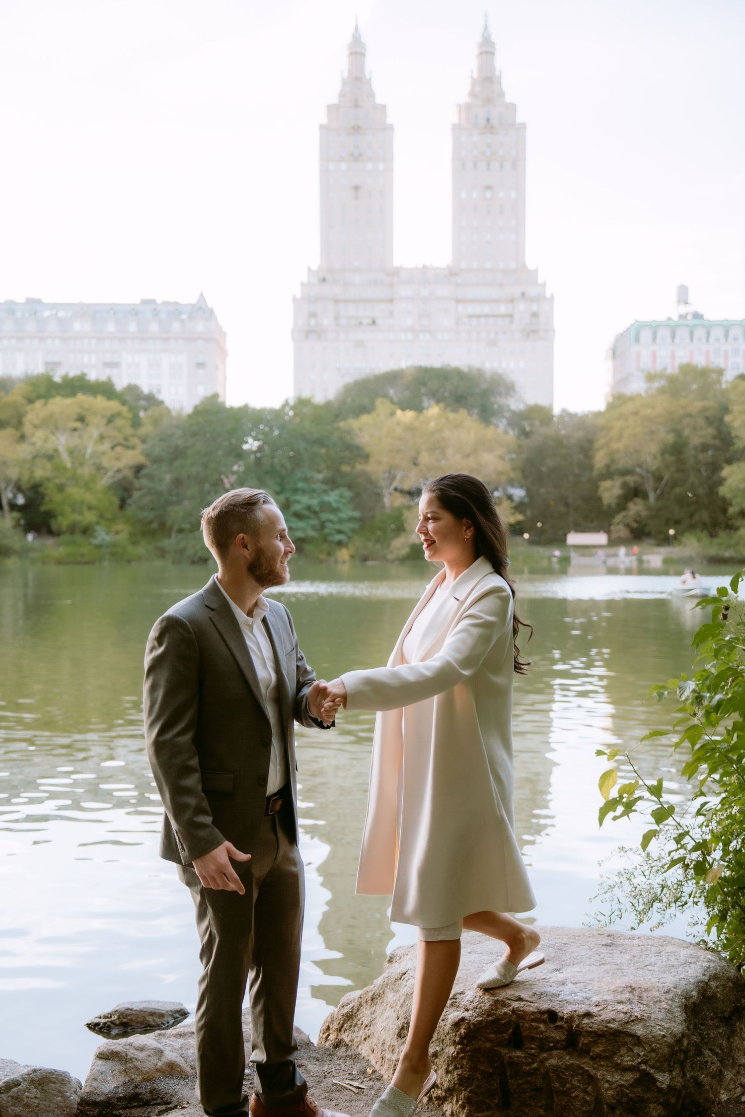 Couple holding hands by a lake in Central Park, NYC, with iconic San Remo building in background.
