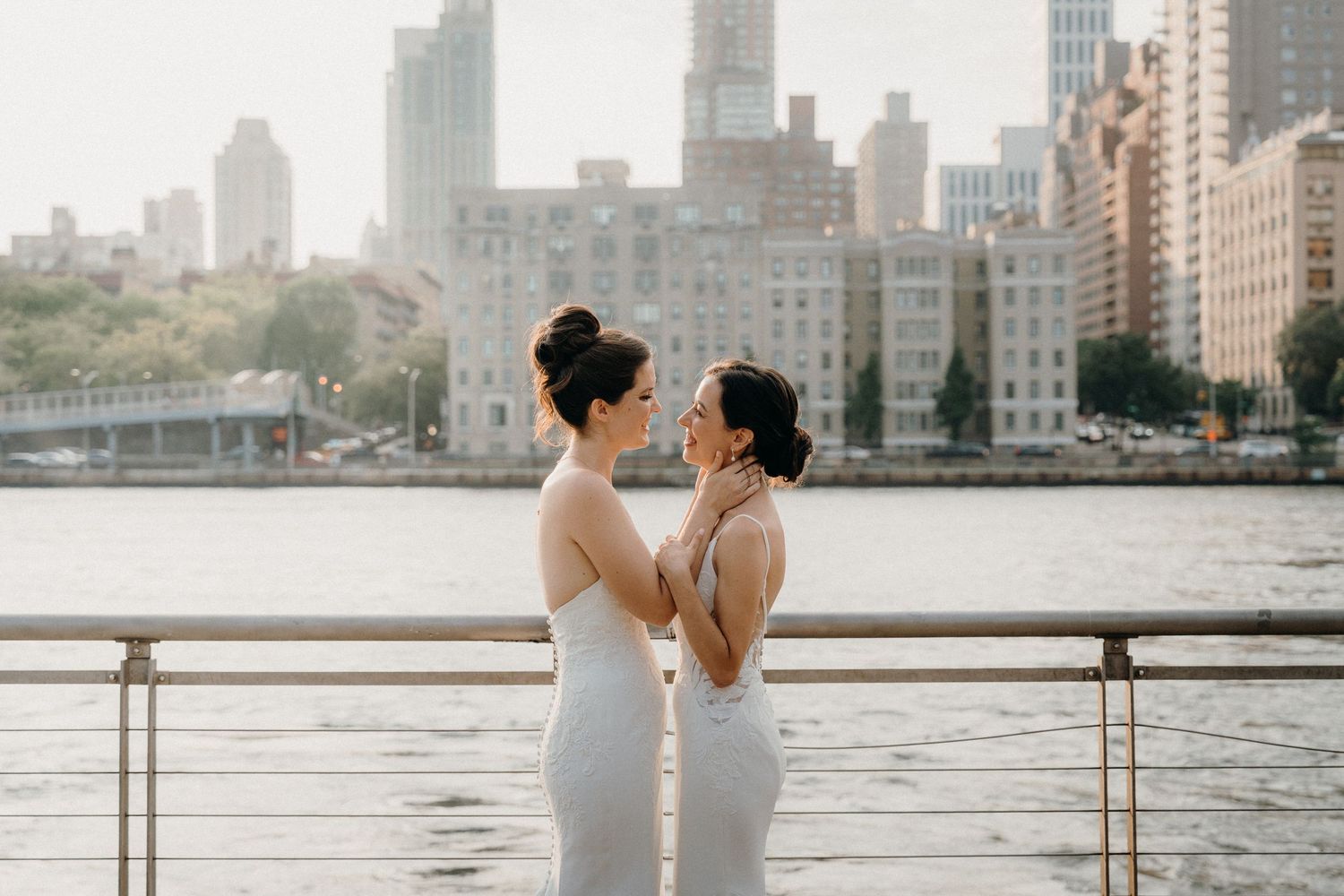 Two brides in white gowns share an intimate moment by the waterfront with NYC skyline backdrop.