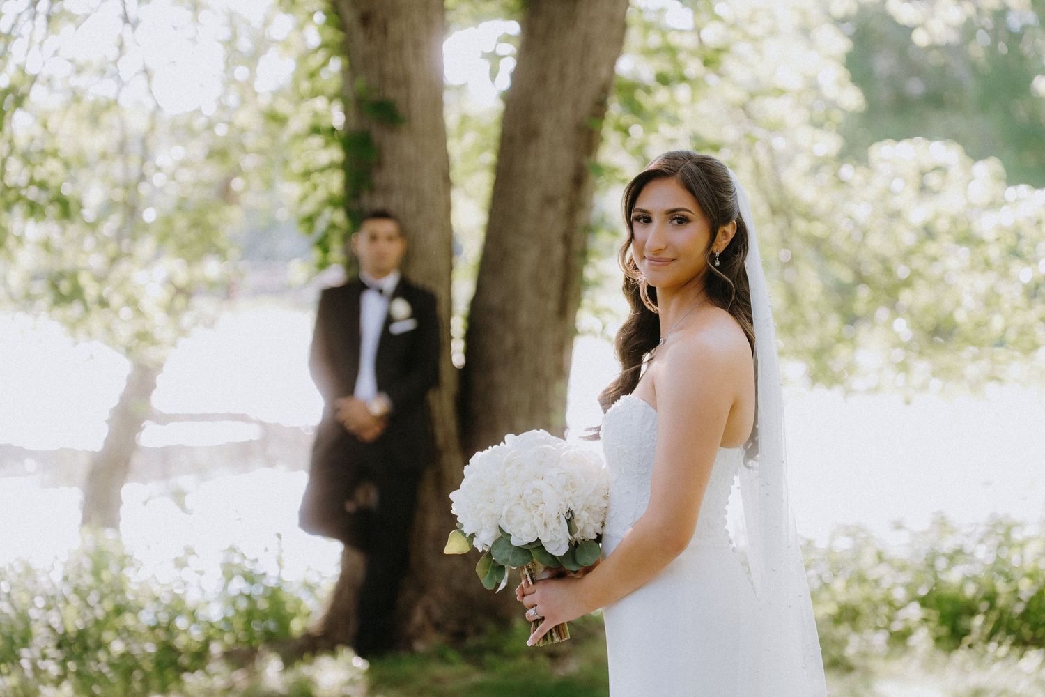 Bride in white gown holds white rose bouquet outdoors, groom in tuxedo stands near tree by waterfront.