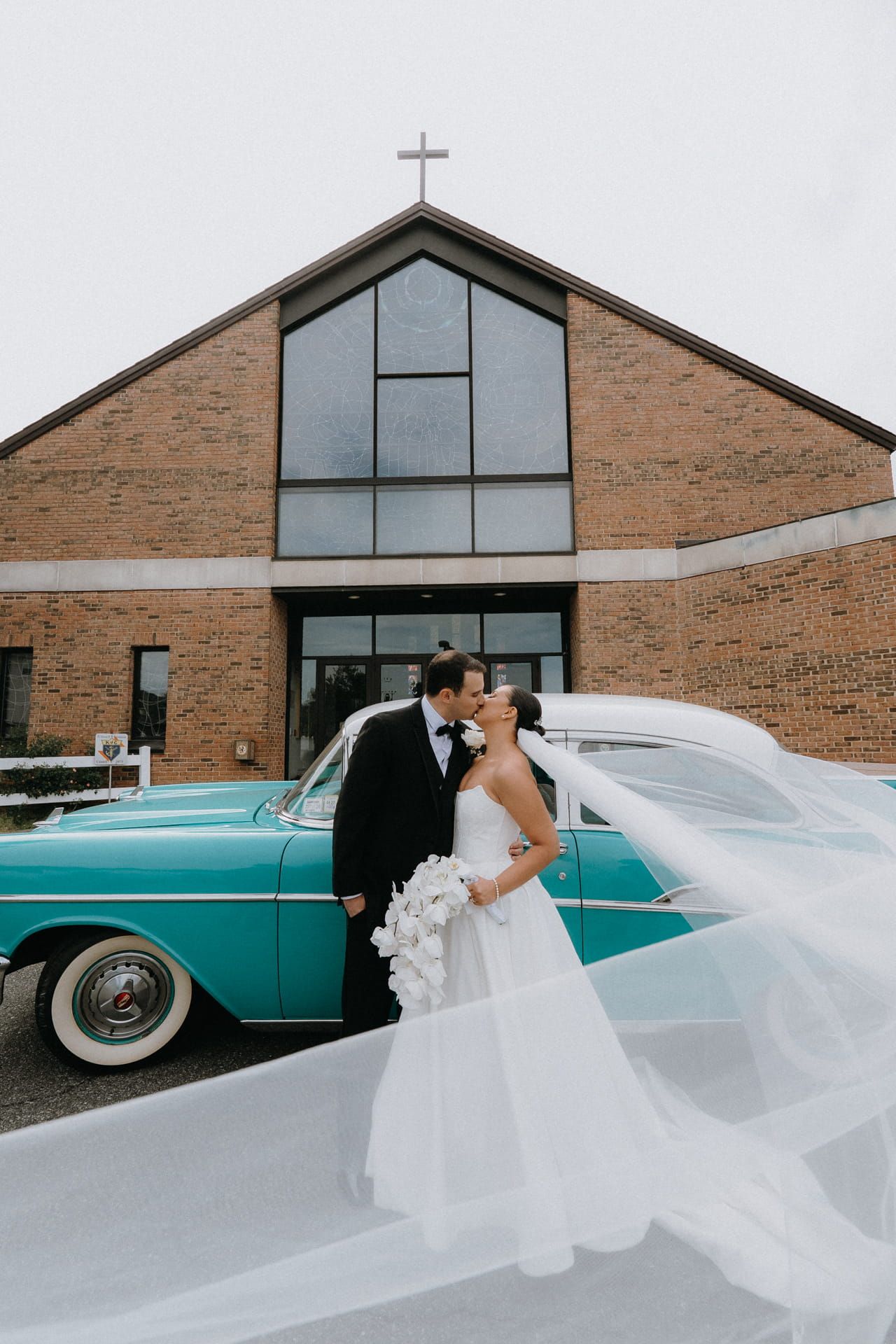 Bride and groom kissing in front of a brick church with a teal vintage car, bride's veil flowing in the wind.