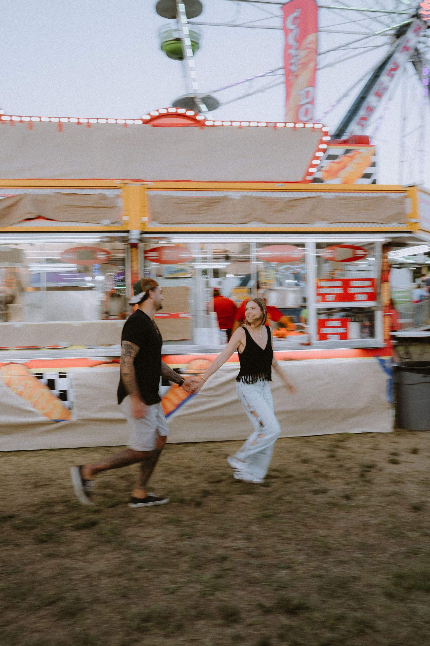 A couple holds hands running past a colorful carnival game booth with a Ferris wheel in the background.