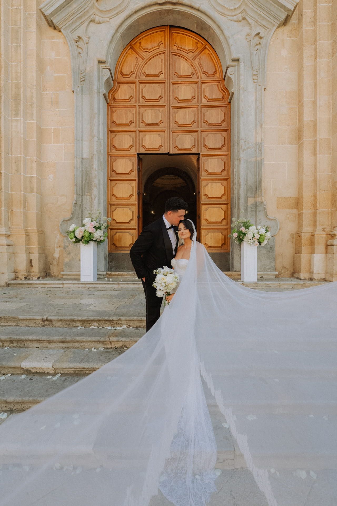 Bride and groom share a romantic moment outside ornate church doors, with flowing white veil billowing in the breeze.