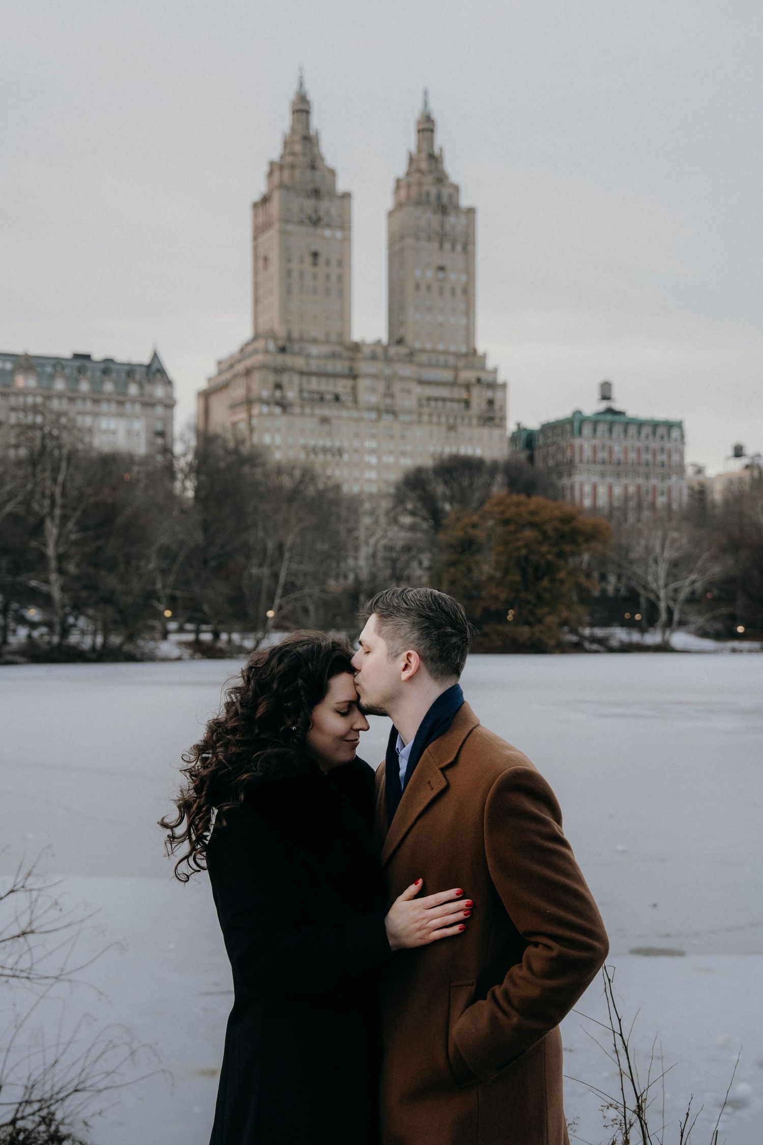 Couple sharing a romantic kiss in Central Park with frozen lake and San Remo building backdrop in winter.