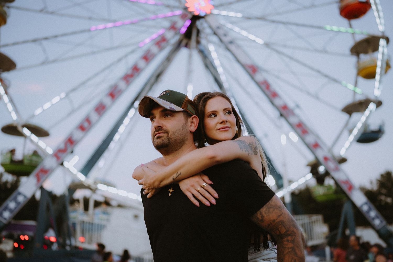 Couple embracing in front of a lit Ferris wheel at an amusement park during dusk.