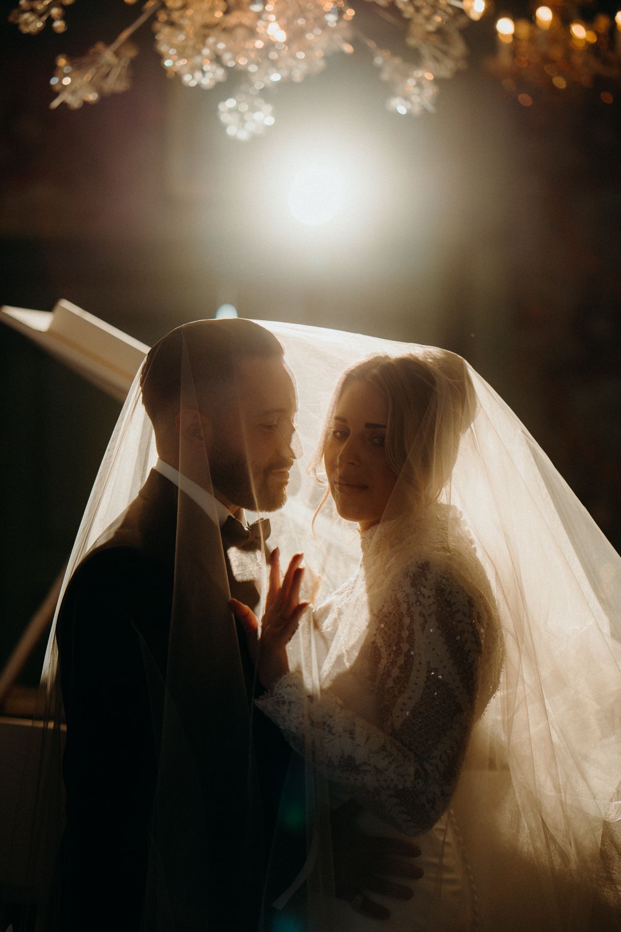 Bride and groom share romantic moment under flowing veil, bathed in warm golden light at elegant wedding ceremony.