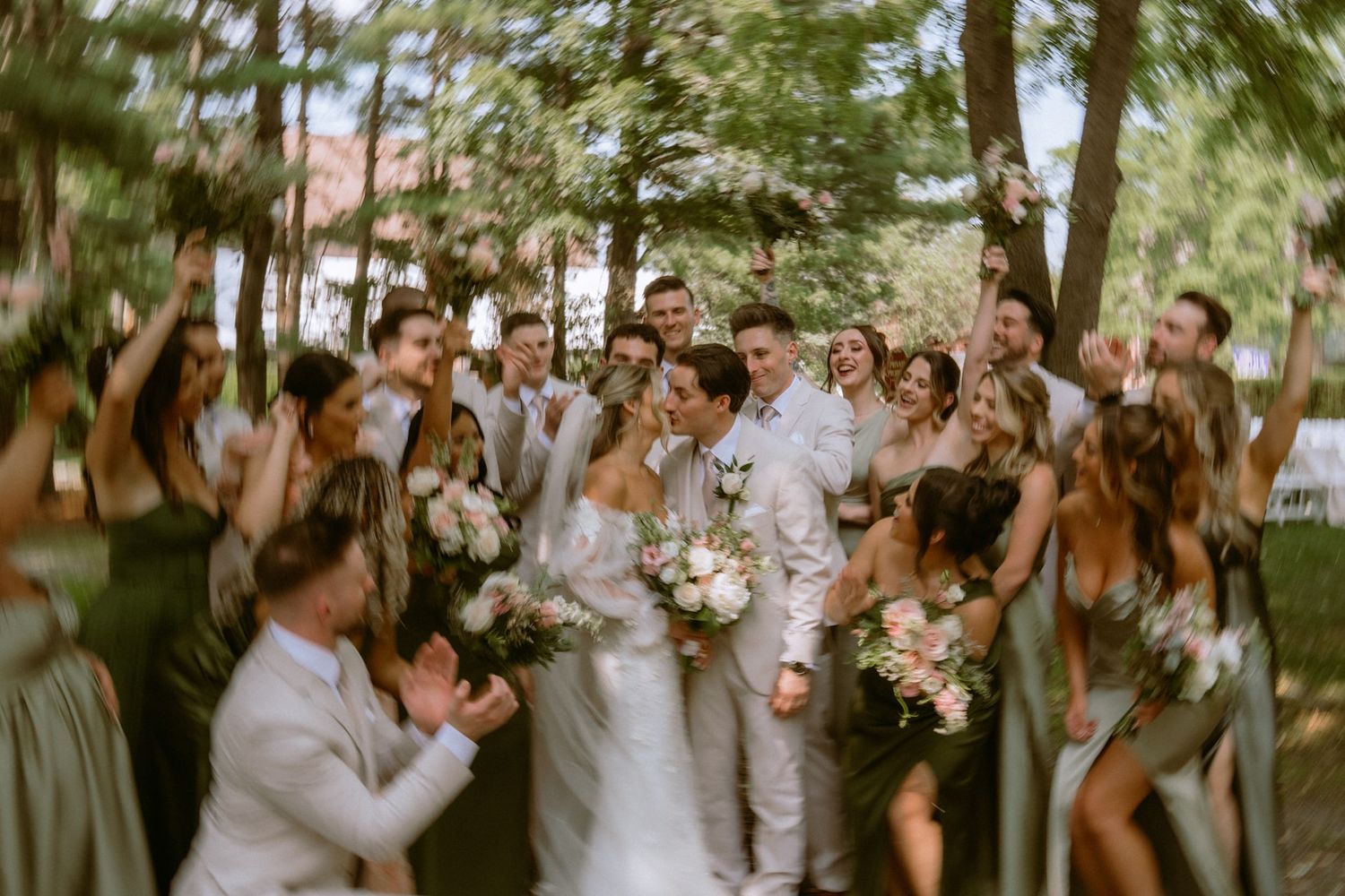 Joyful wedding party celebrating with bride and groom outdoors, surrounded by bridesmaids in sage green dresses and groomsmen.
