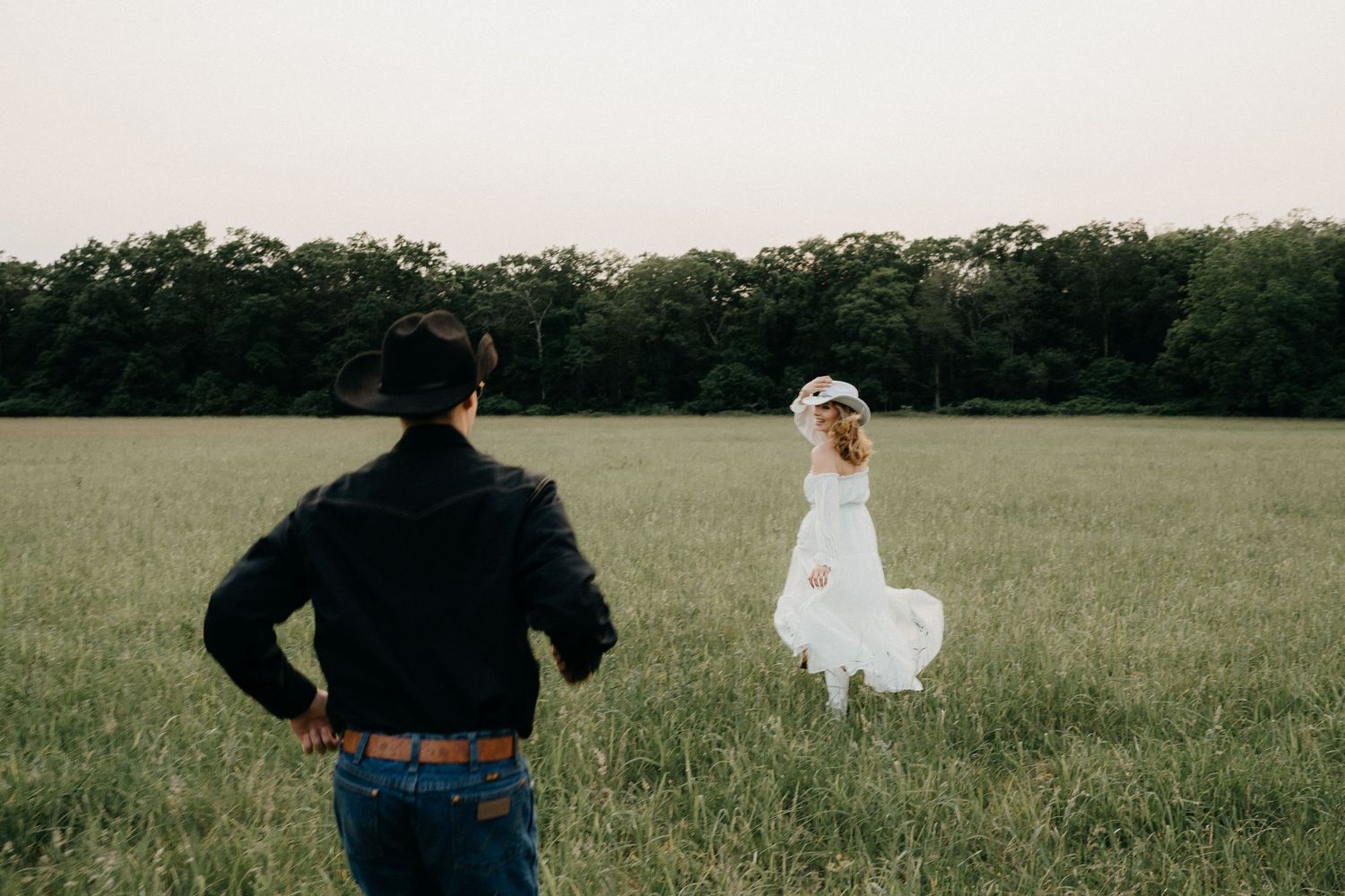 A couple in a field, man in cowboy hat and jacket, woman in white dress and floral crown, at dusk.