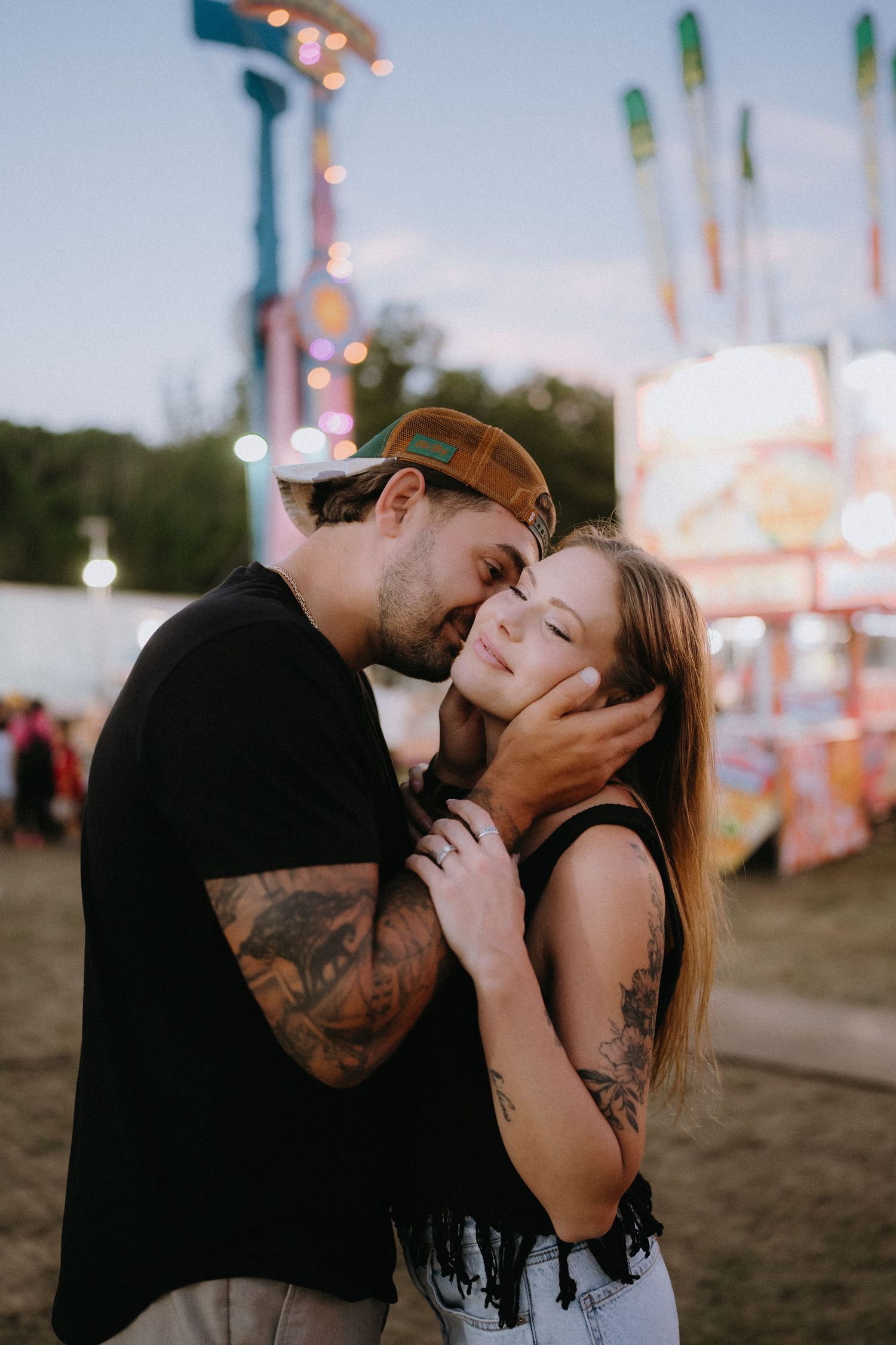 Romantic couple sharing a tender moment at a carnival, man with tattoos gently holding woman's face at dusk.