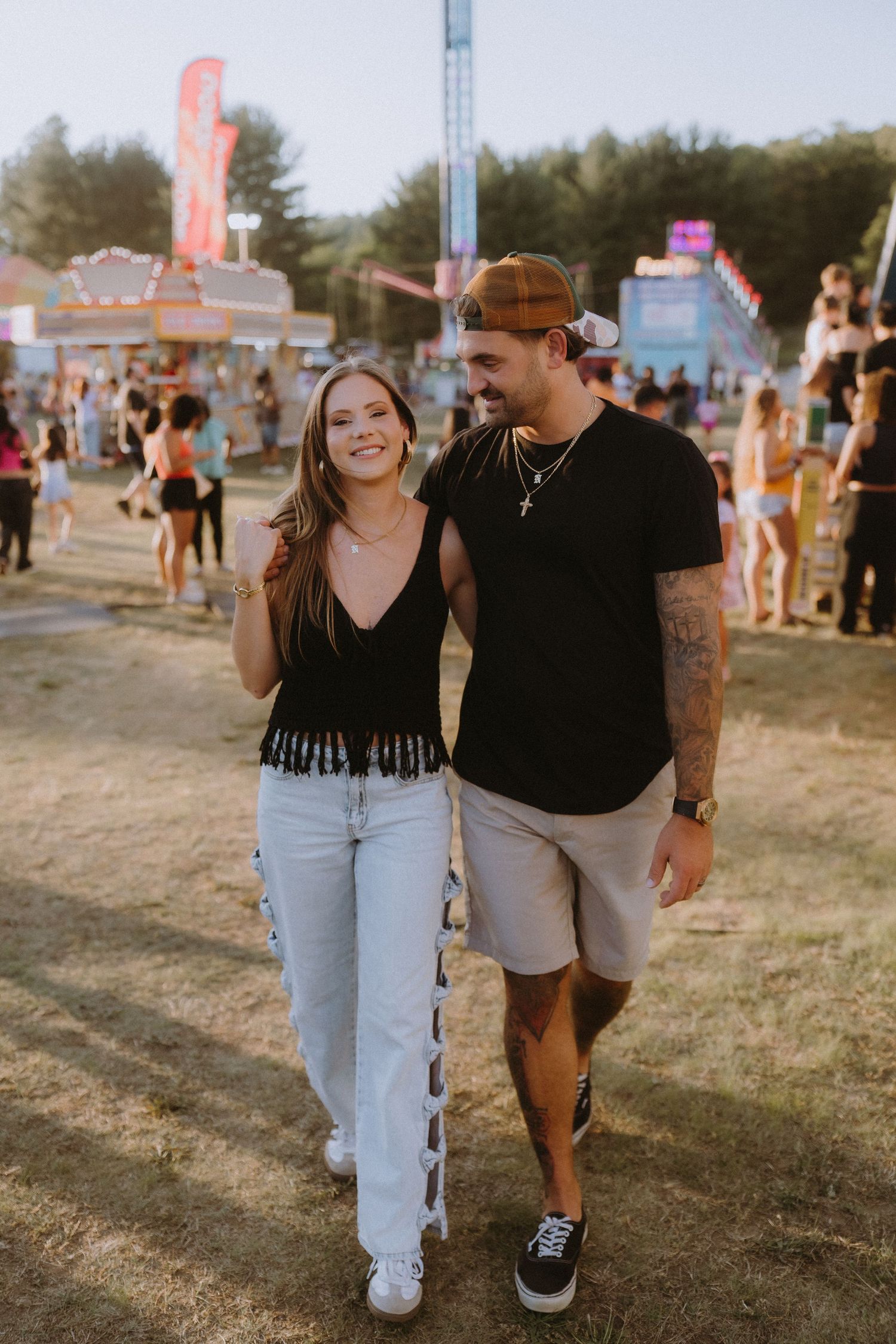 Happy couple walking together at an outdoor festival, woman in black fringe top and jeans, man in black t-shirt and shorts.