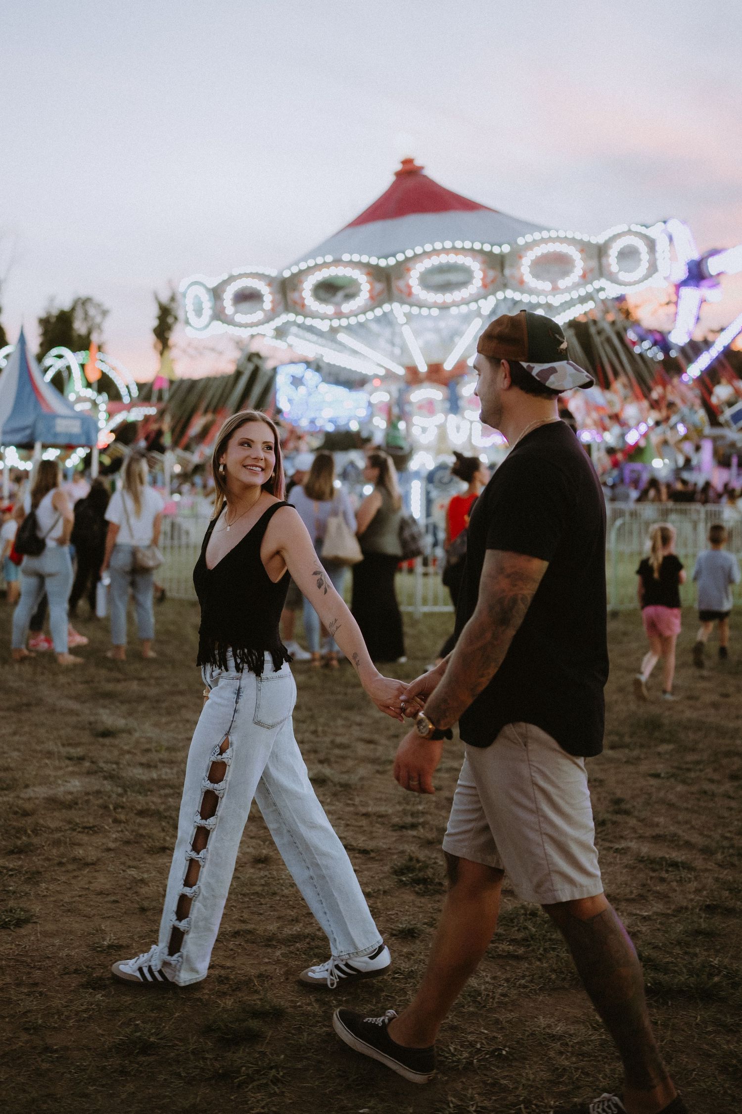 A couple holds hands and smiles while walking at a colorful carnival at dusk with a lit carousel in the background.