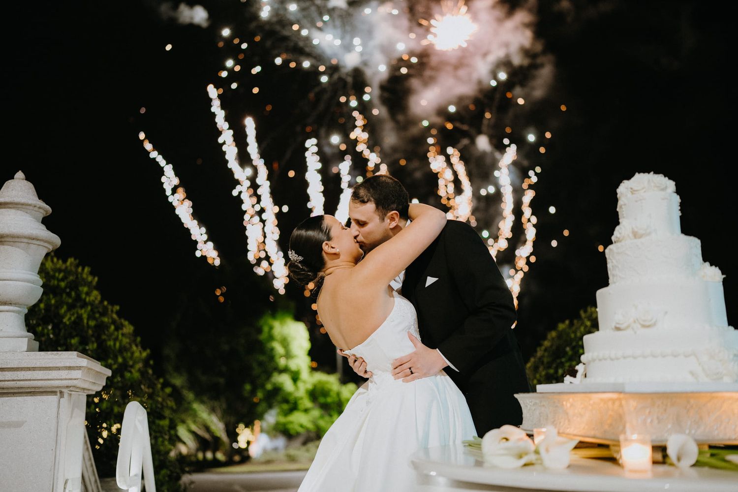 Bride and groom share a romantic kiss in front of stunning fireworks display beside a white wedding cake at night.