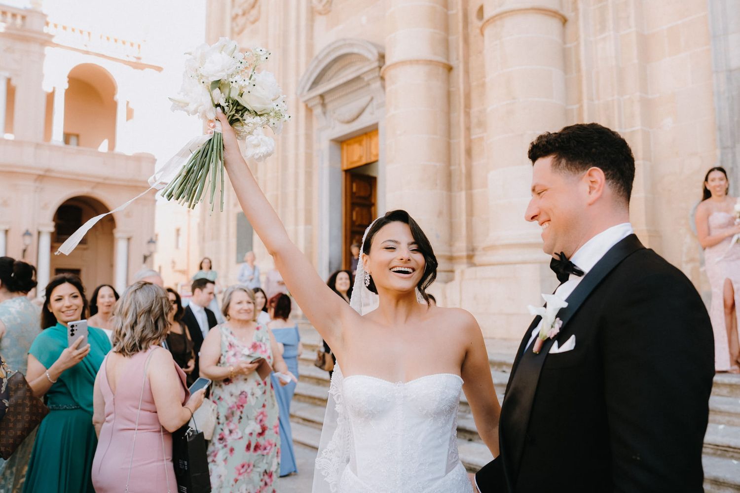 Bride raises bouquet joyfully outside ornate church as groom in tuxedo smiles beside her after wedding ceremony.