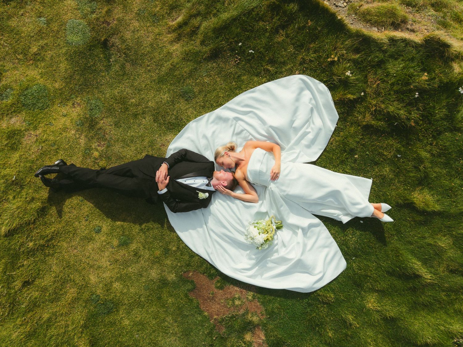 Aerial view of newlyweds lying on green grass, bride in white gown with flowing train, groom in black suit, holding bouquet.