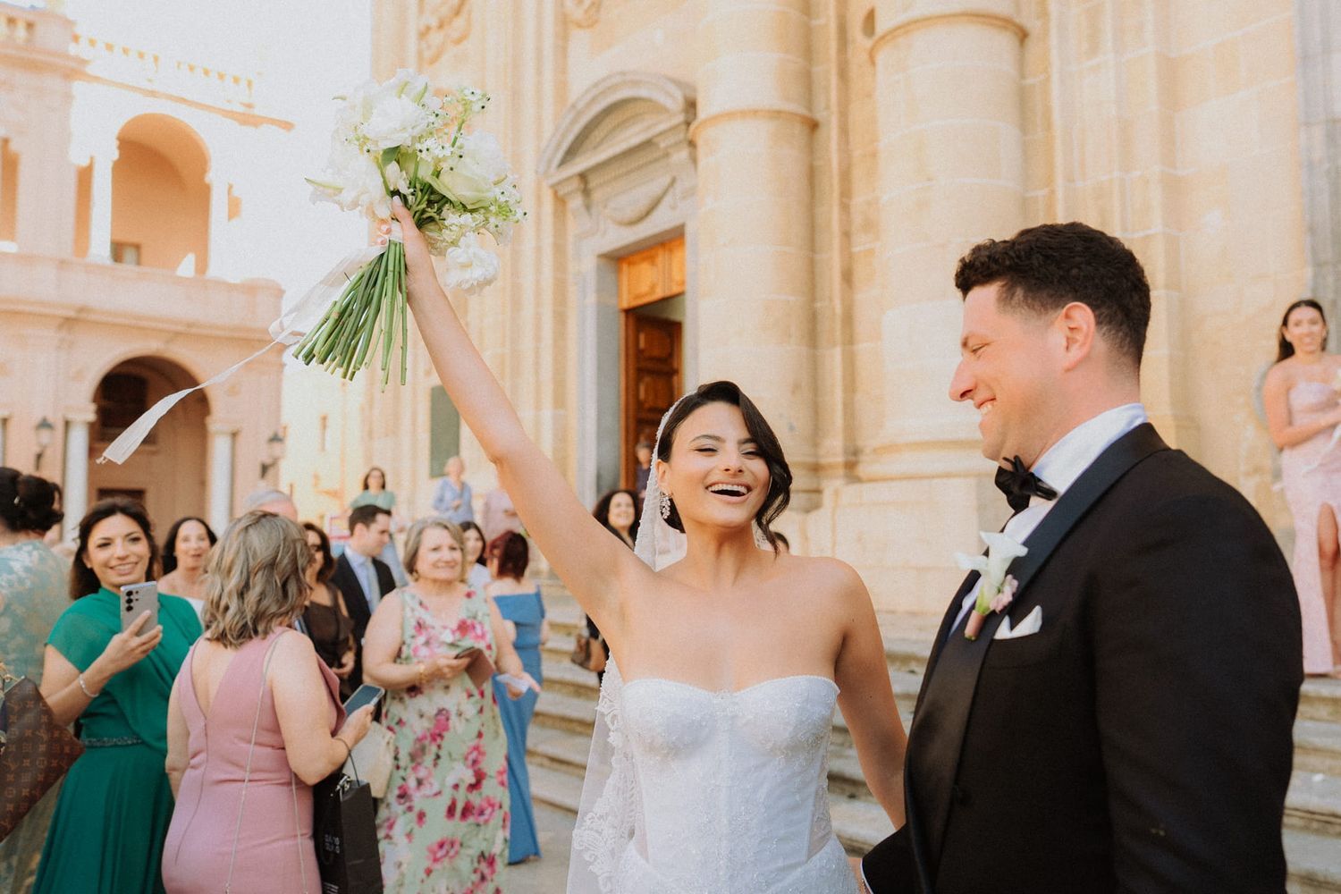 Joyful bride raises bouquet outside ornate church as groom smiles beside her, guests celebrating in background.
