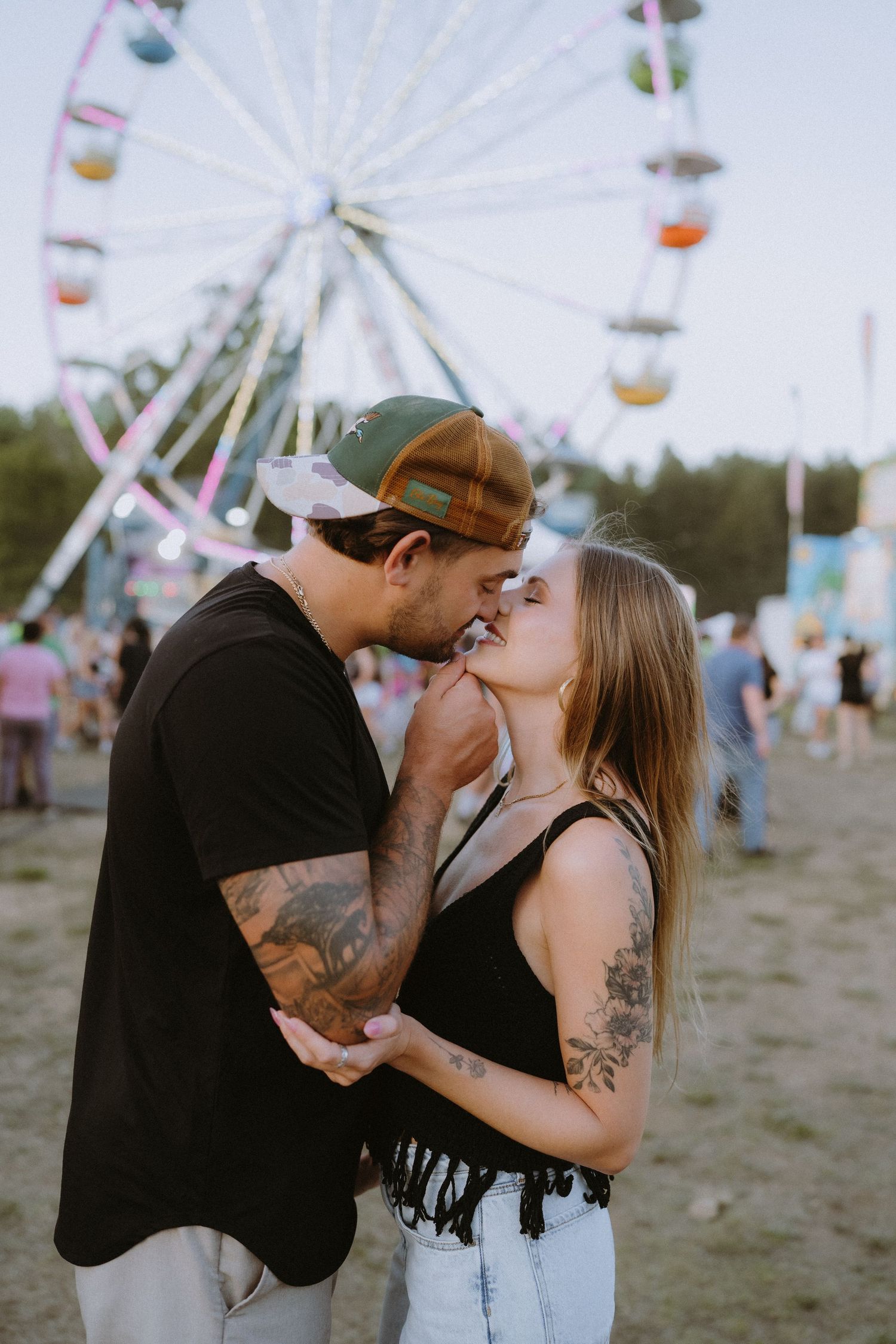 Couple kissing at outdoor festival with ferris wheel backdrop, both showing tattoos, woman in black top.