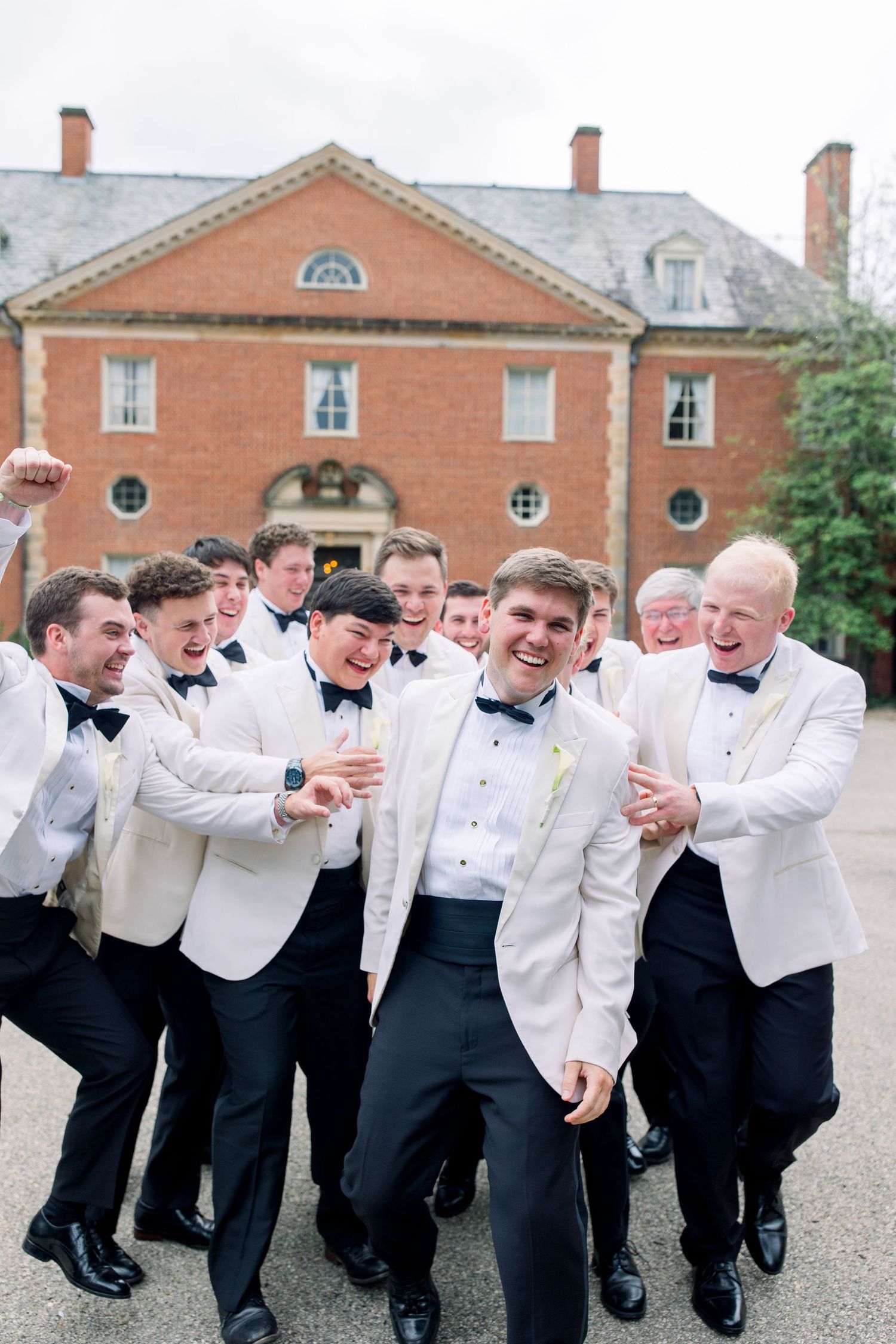Groom and groomsmen in white tuxedo jackets and black bow ties laughing together outside a brick mansion.