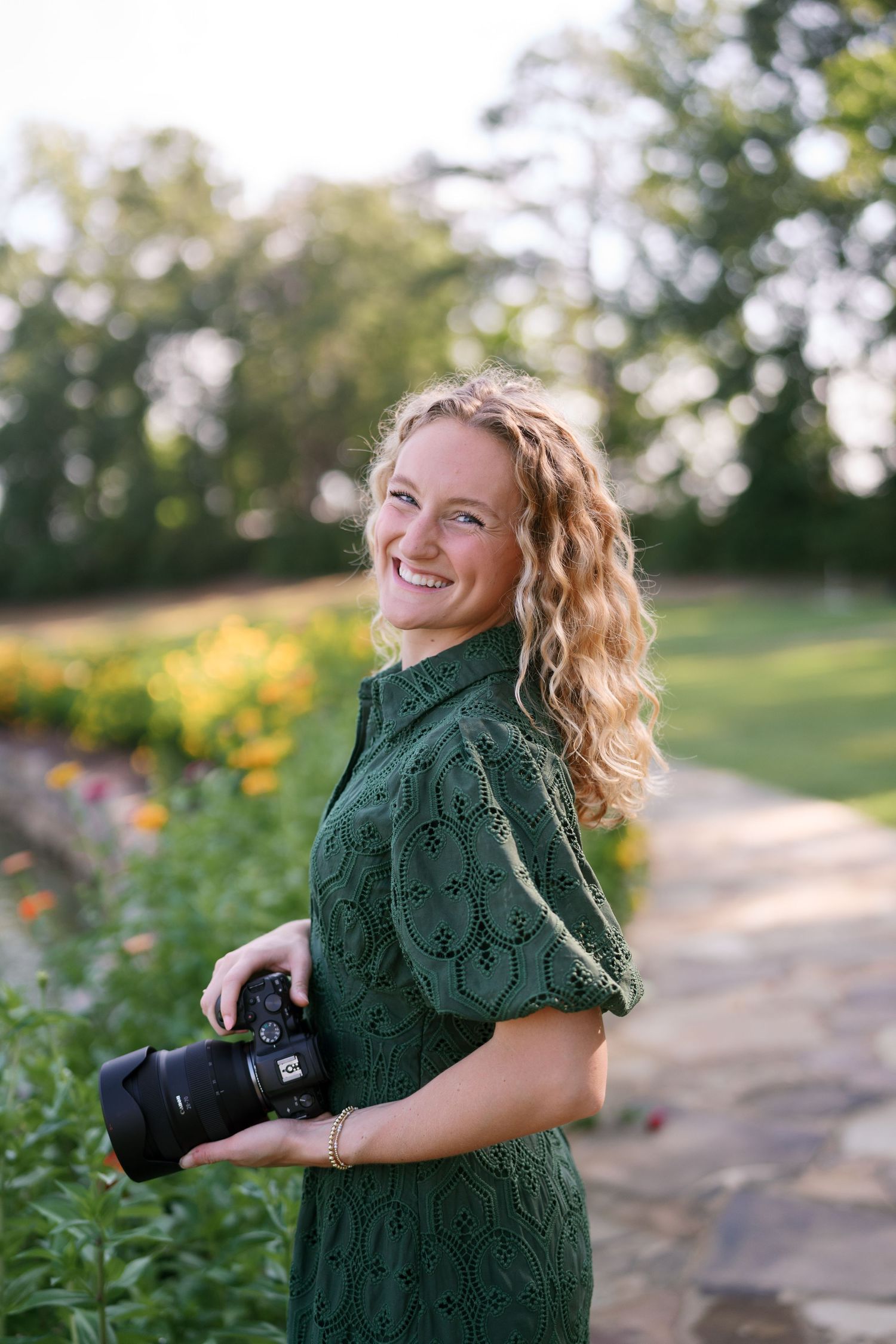 Smiling woman with curly hair in green lace dress holding a camera outdoors in a garden setting.