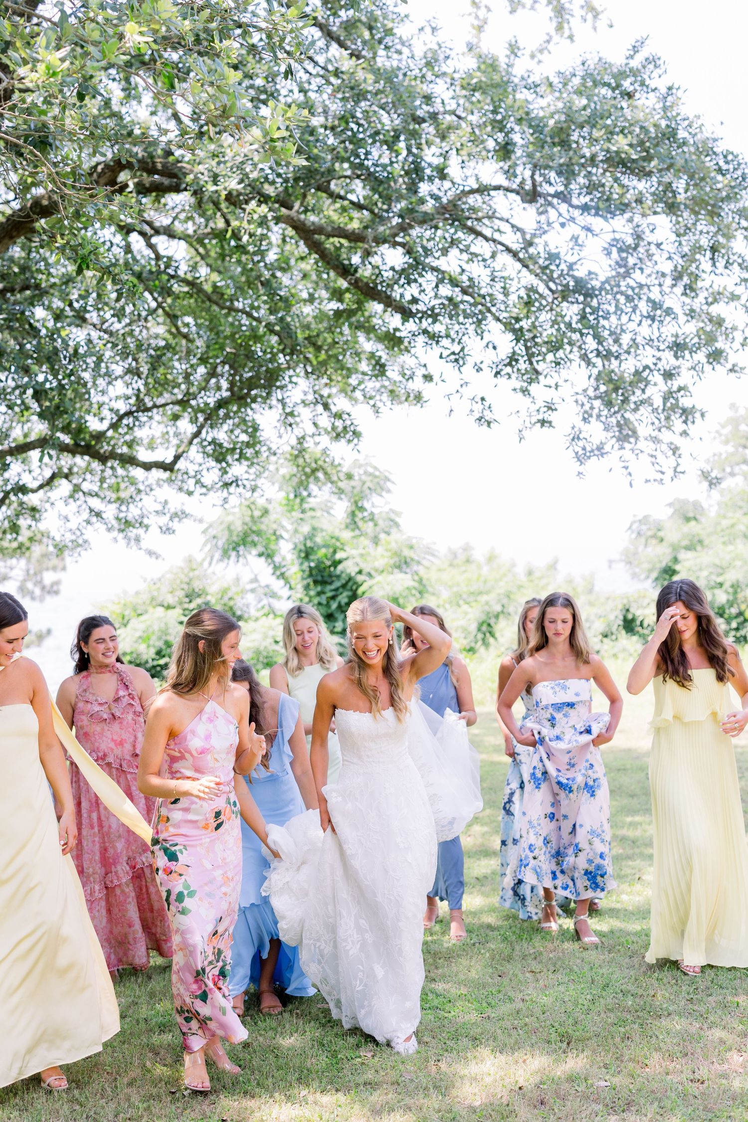 Wedding party in pastel dresses poses under a large oak tree on a sunny day.