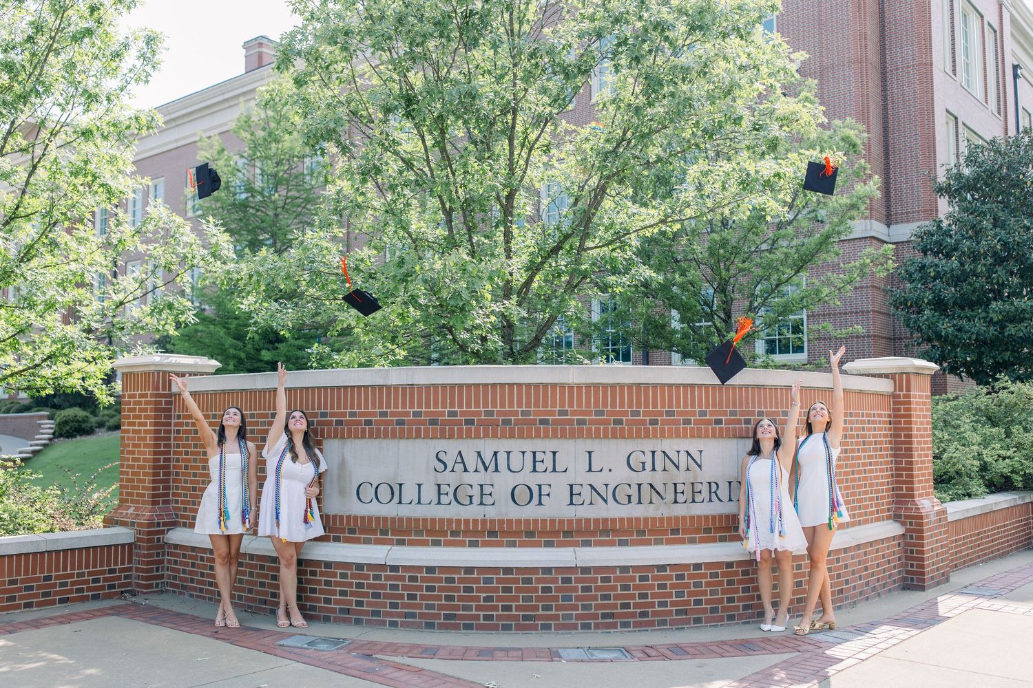 Two graduates in white dresses toss caps at Samuel L. Ginn College of Engineering sign at Auburn University.