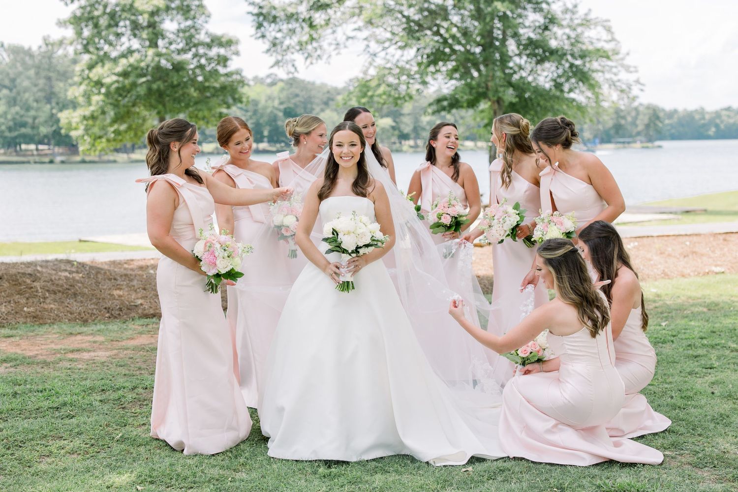 Bride in white ball gown surrounded by bridesmaids in blush pink dresses outdoors near a lake.