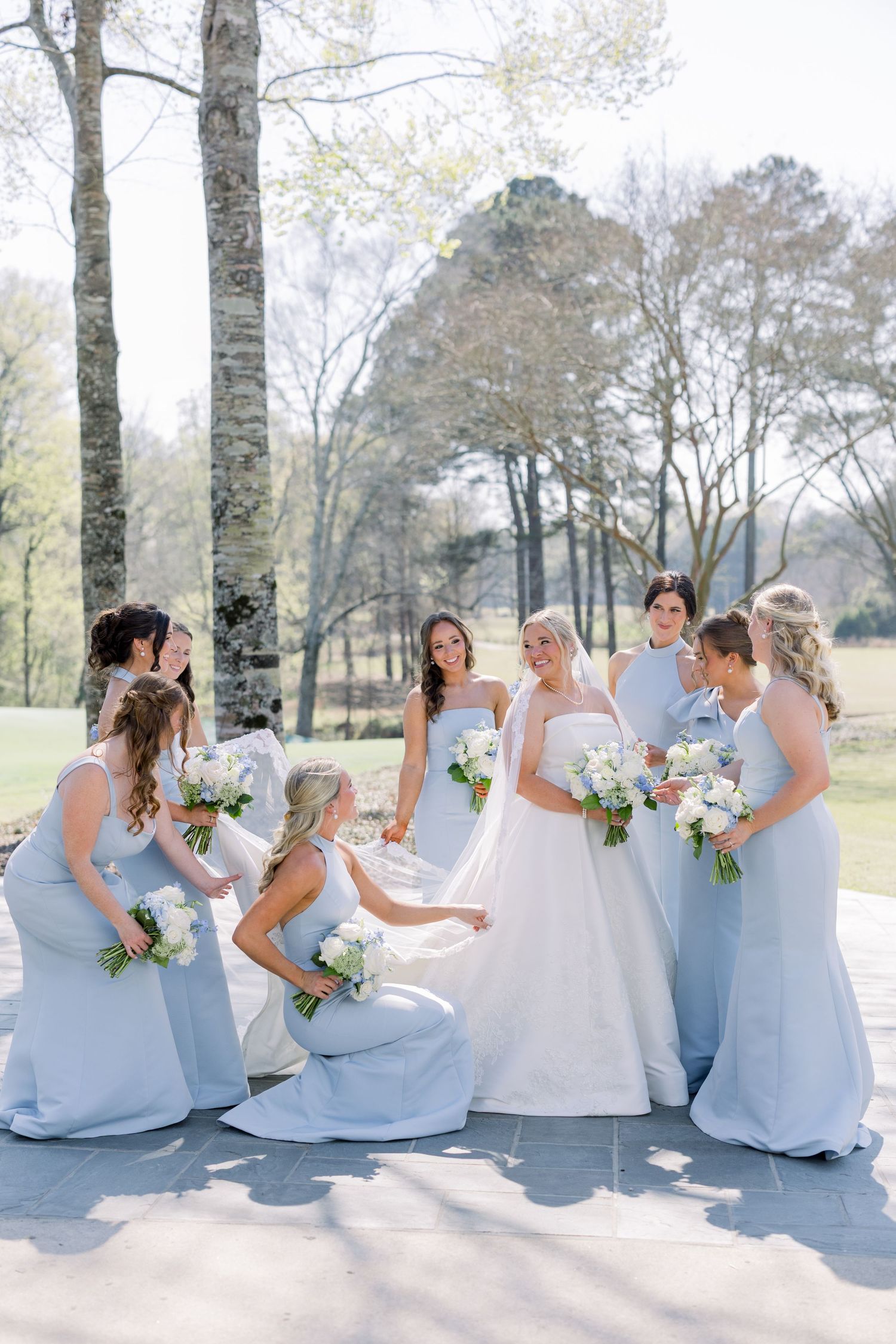Bride in white gown surrounded by bridesmaids in light blue dresses holding bouquets outdoors at a spring wedding.