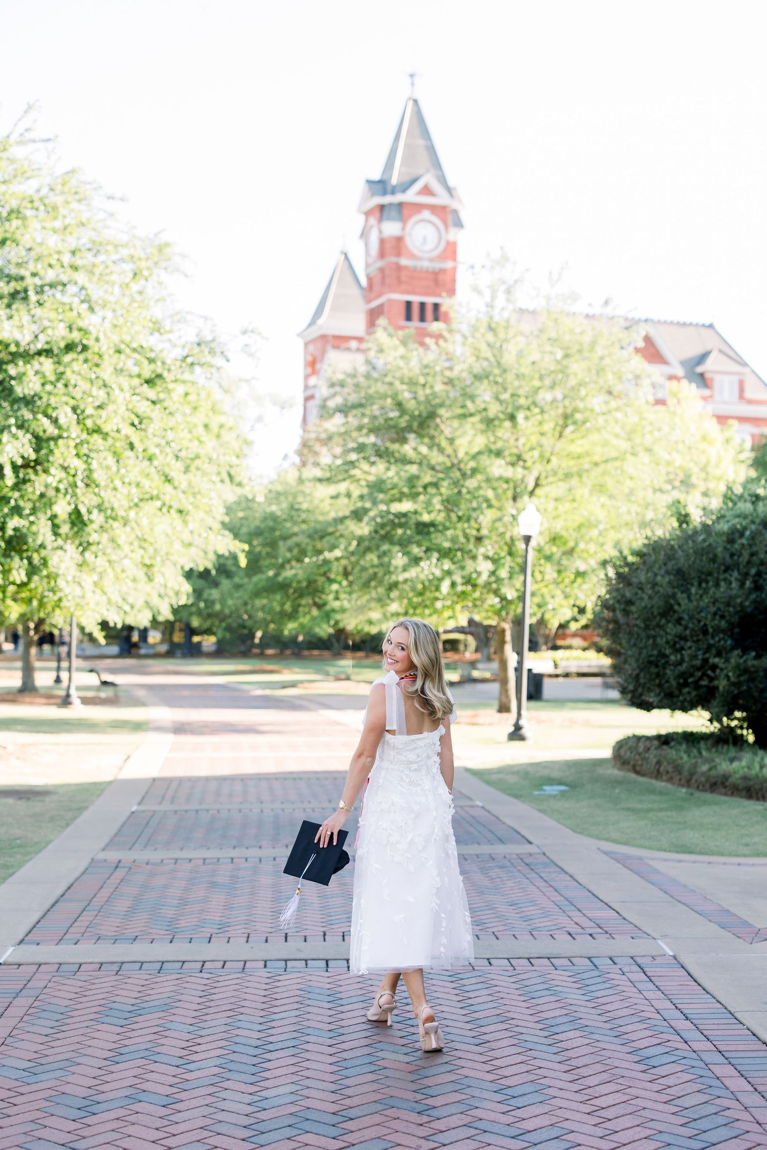 Blonde woman in white midi dress holding clutch on brick plaza near red brick clock tower building.