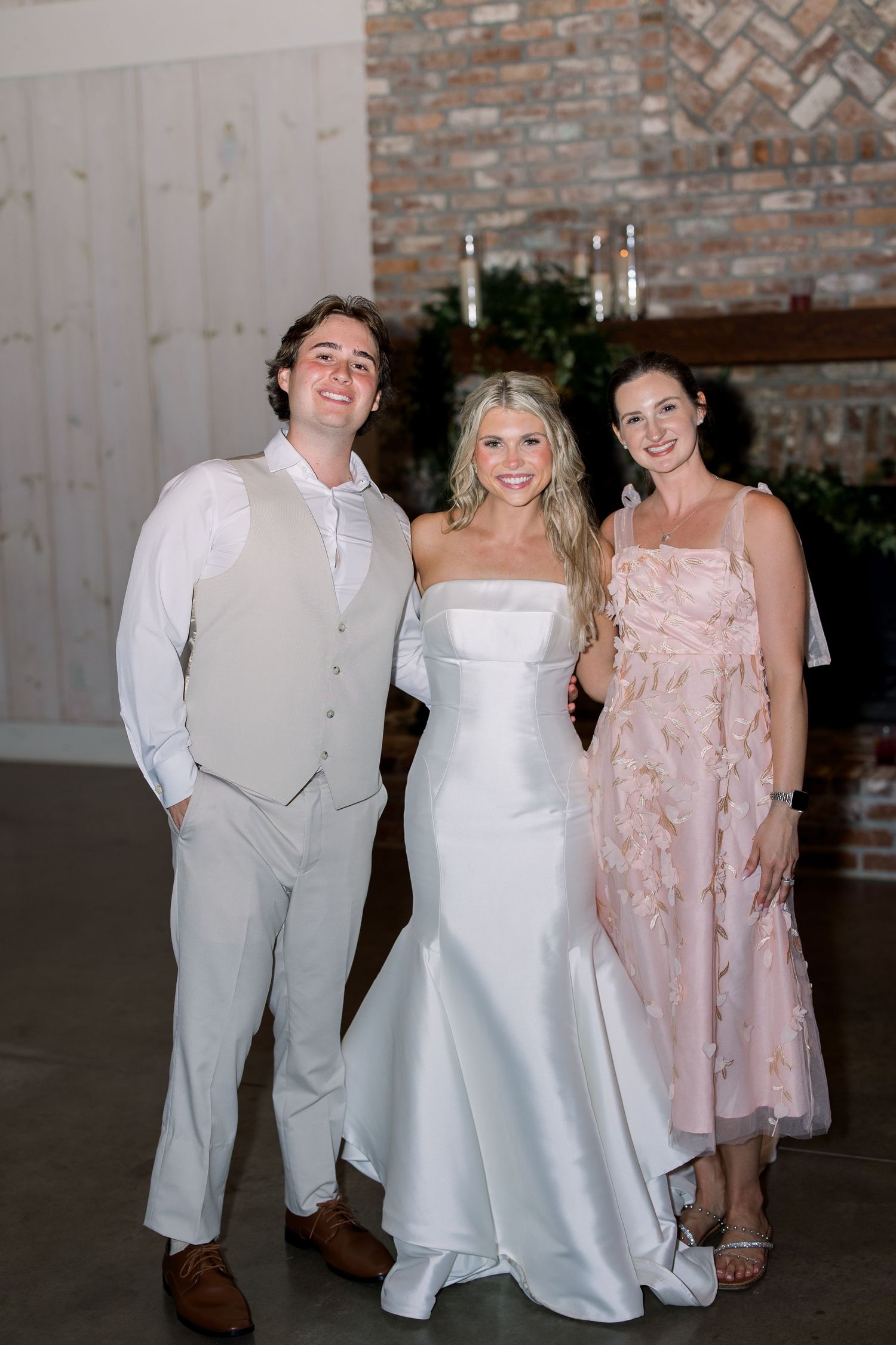 Bride in white gown poses with man in gray vest suit and woman in floral pink dress at elegant wedding venue.