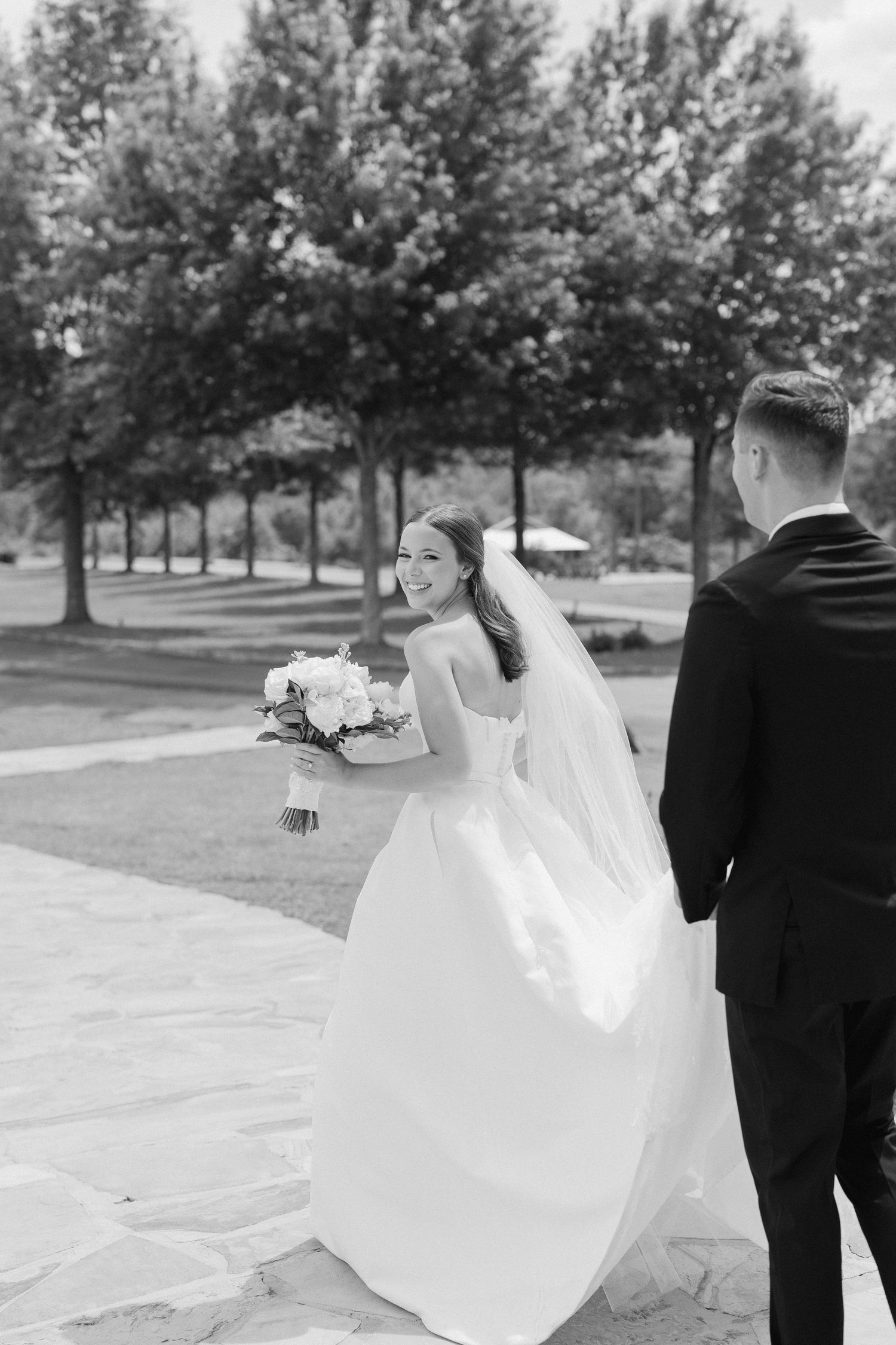 Black and white photo of a smiling bride in a flowing gown holding a bouquet, glancing back at her groom outdoors.