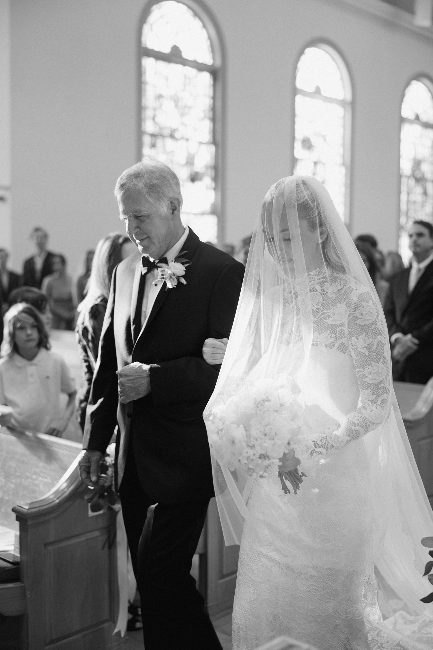 Black and white photo of a bride in lace veil walking down church aisle with her father on wedding day.
