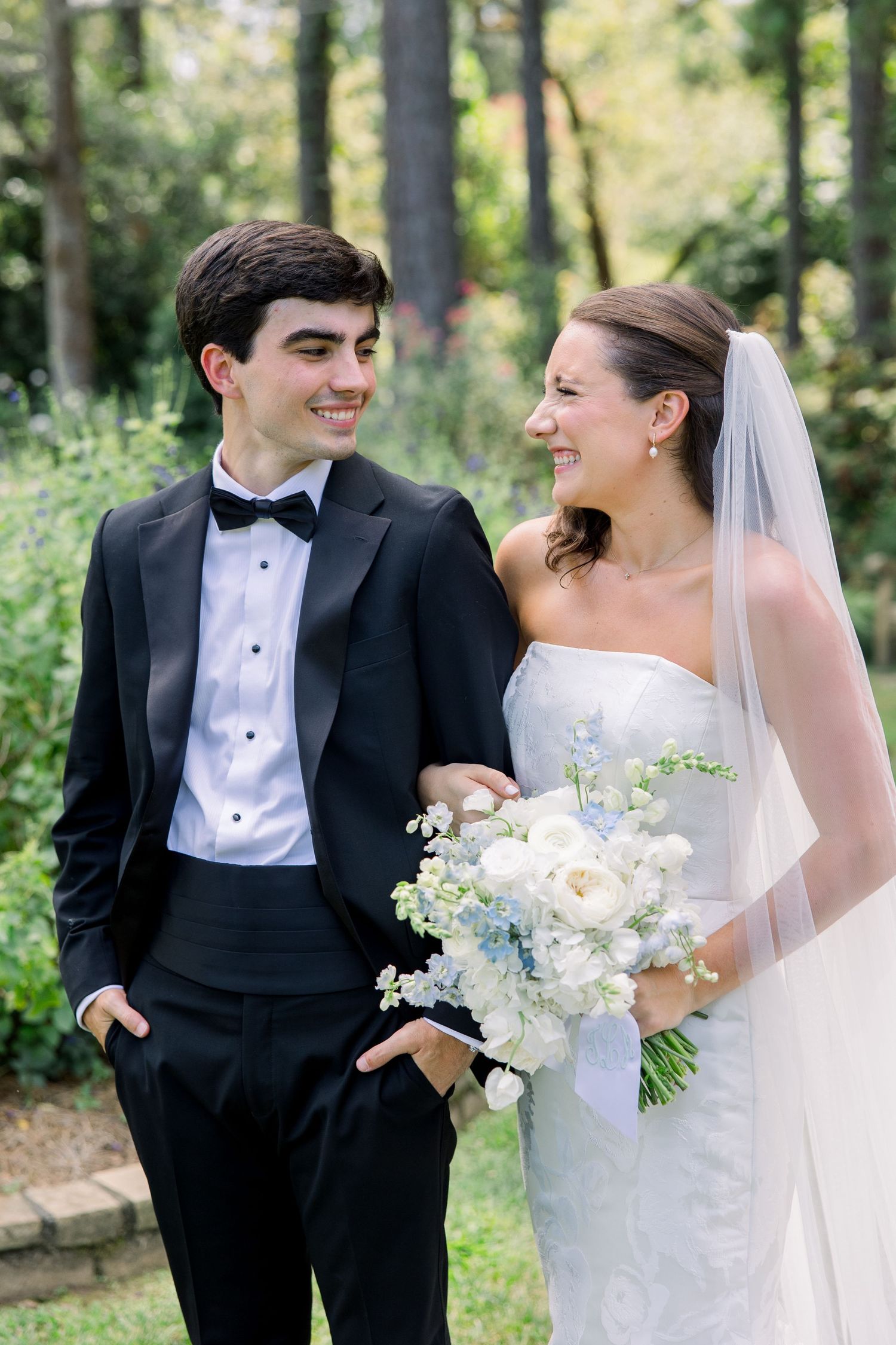A smiling bride in a white gown and veil holds a blue and white bouquet beside a young man in a black tuxedo outdoors.