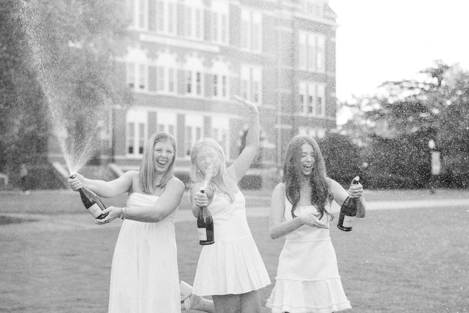 Three people in white dresses celebrate with champagne bottles spraying in a garden setting.