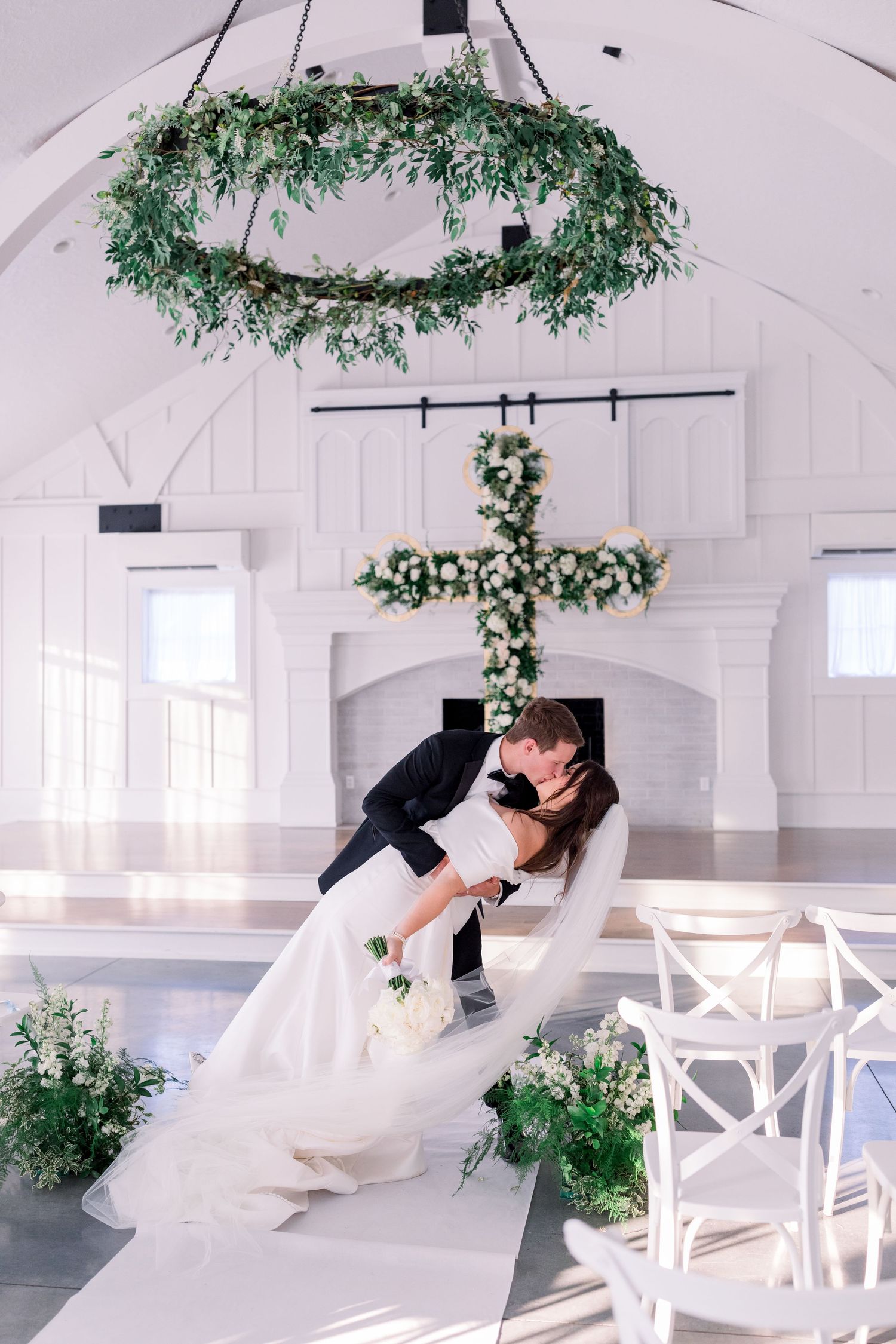Romantic wedding couple sharing a dramatic dip pose in a white chapel decorated with floral cross and greenery.