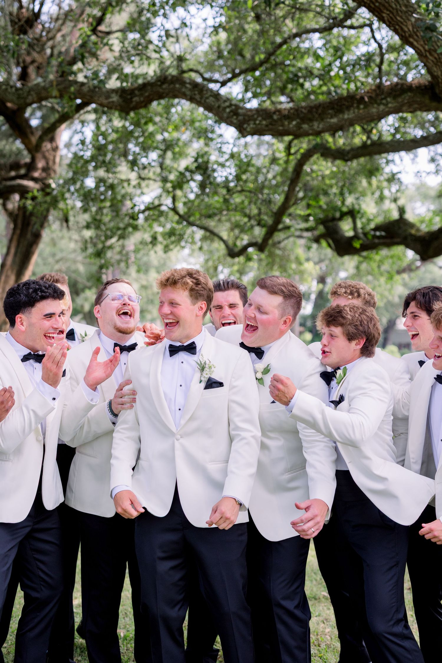 Groomsmen in white tuxedo jackets share a laugh together under a large oak tree.