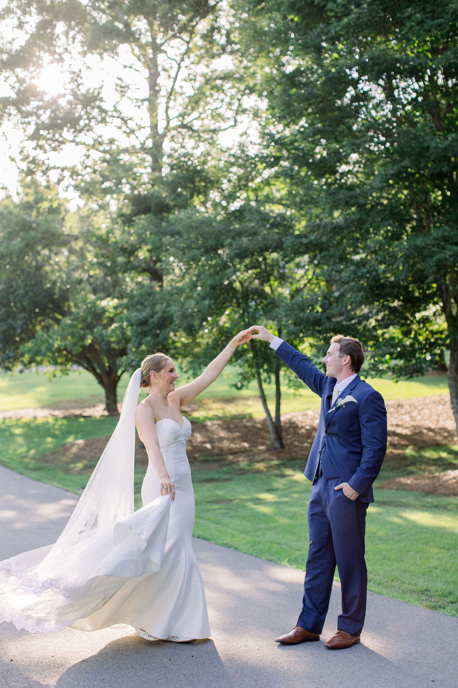 Bride and groom dancing outdoors in lush green park, bride in white gown with veil, groom in navy suit.