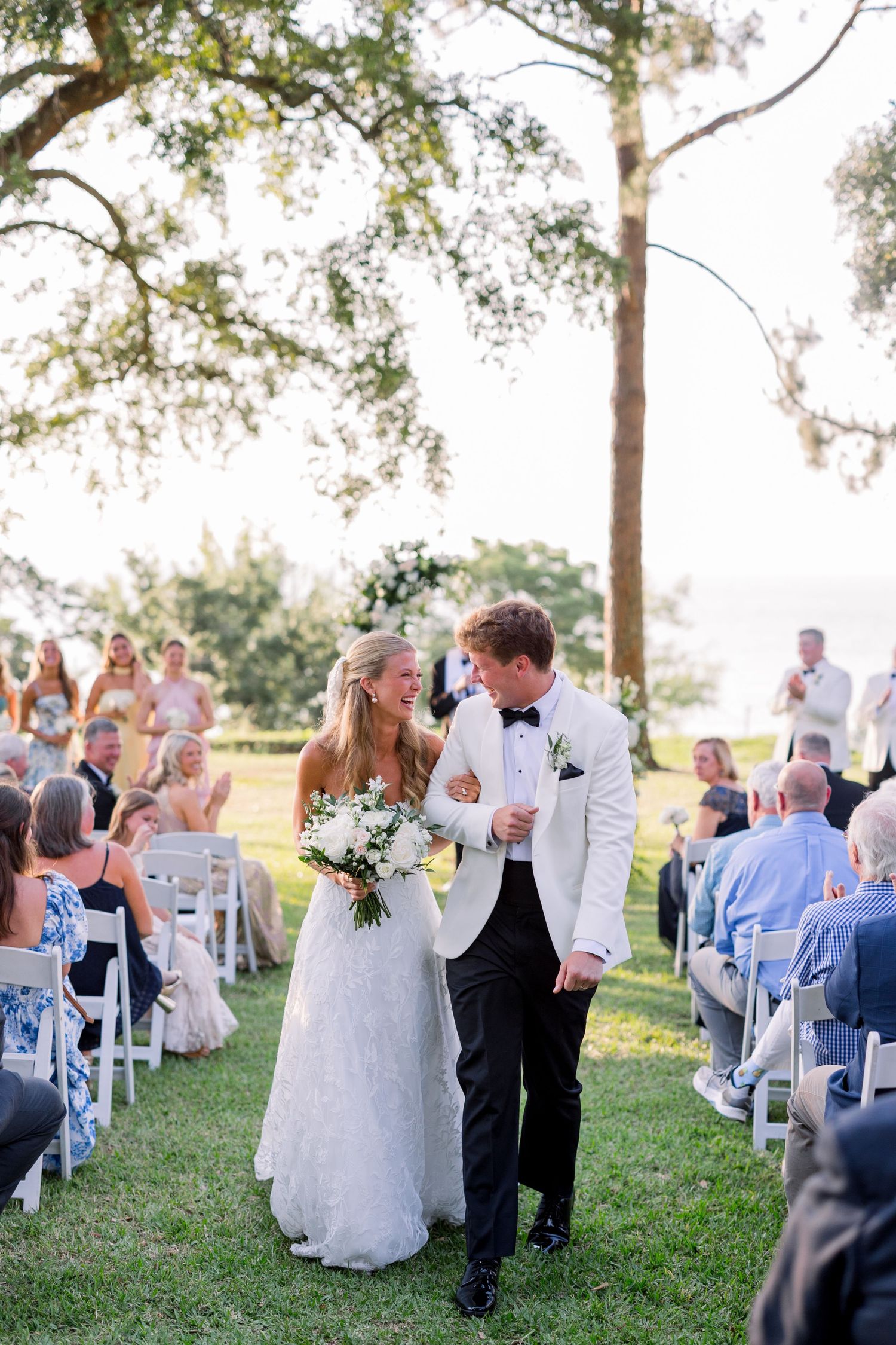Bride and groom walk down outdoor aisle after wedding ceremony, surrounded by seated guests on a sunny day.