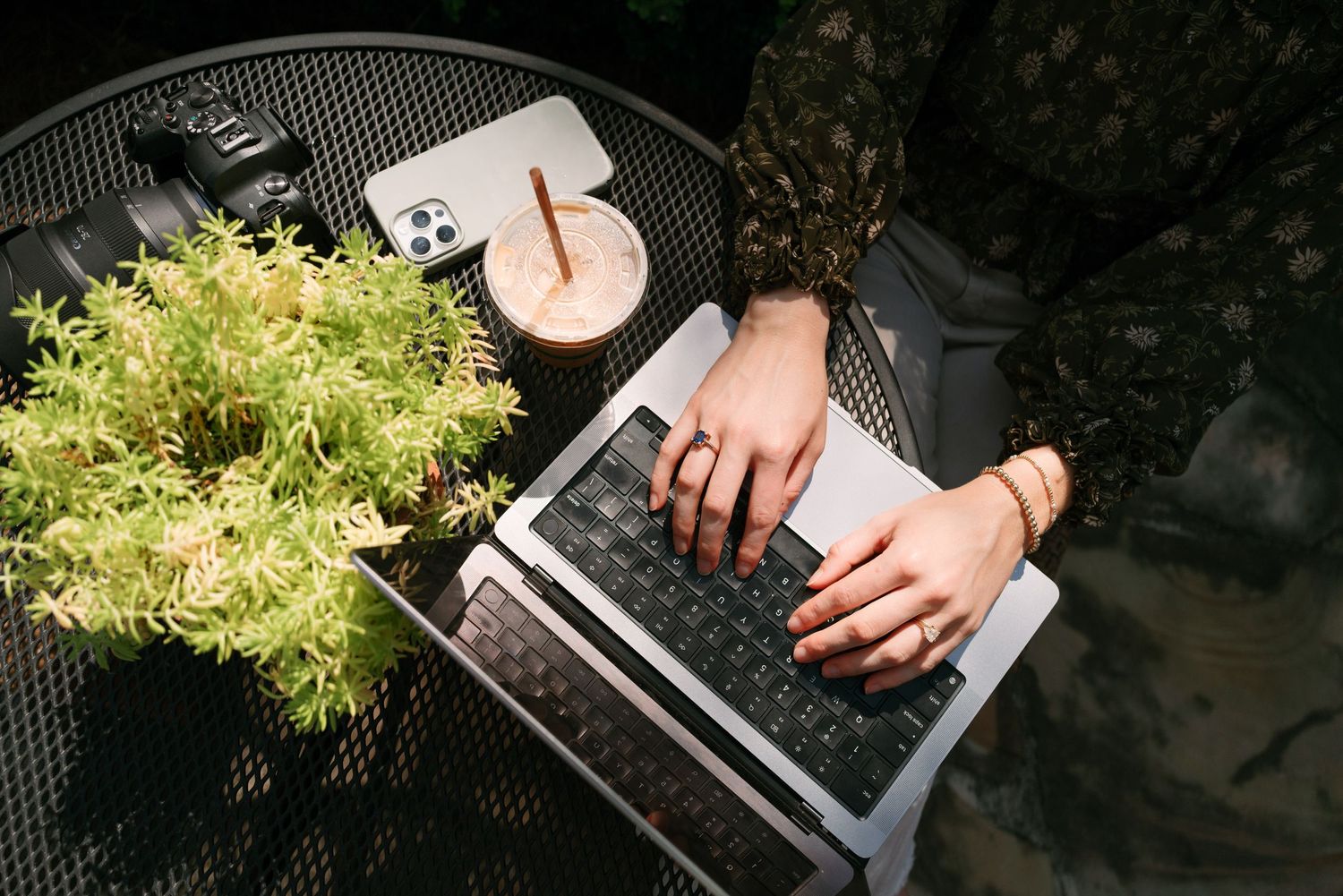 Person typing on a laptop at an outdoor metal table with a smoothie, smartphone, and lush green plant in sunlight.