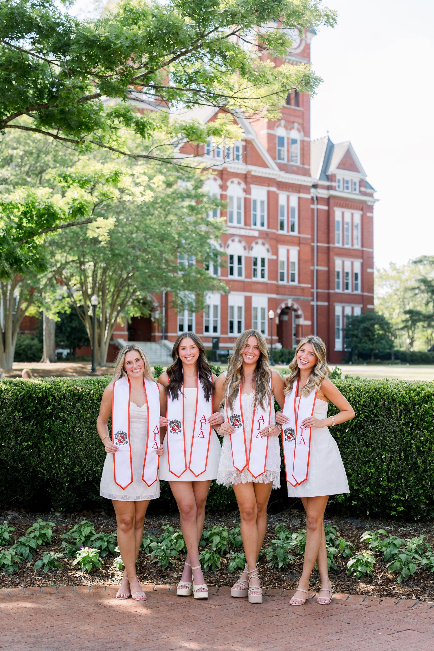 Four college graduates in white dresses and red graduation stoles pose proudly in front of a brick university building.