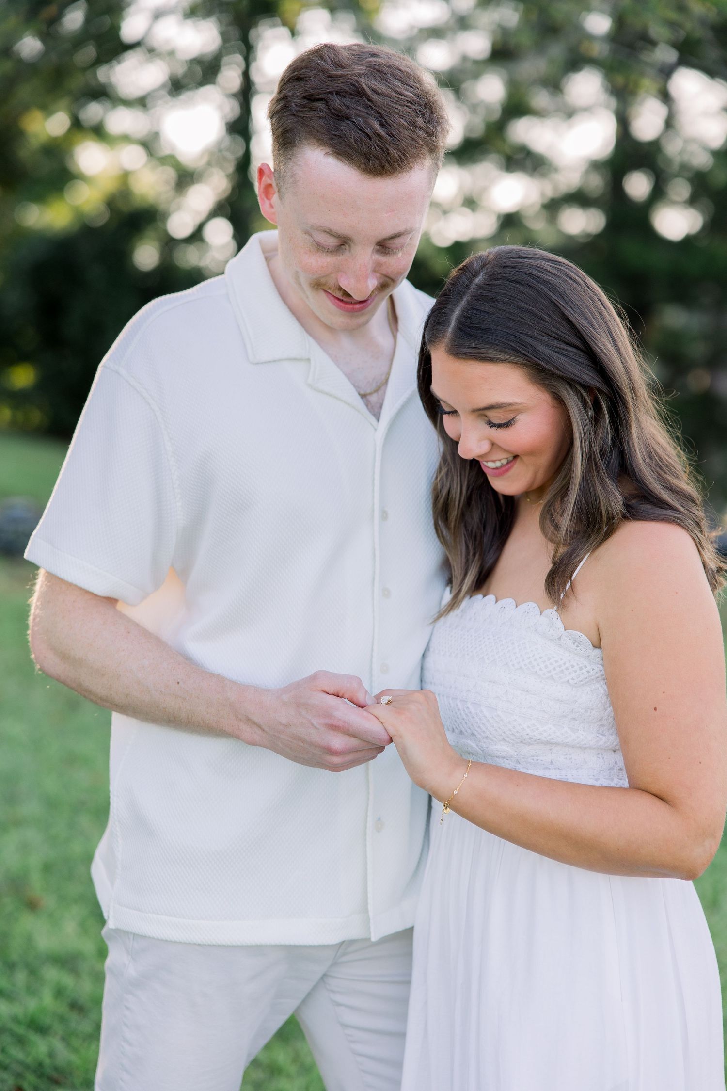 Smiling couple in white outfits looking down at an engagement ring outdoors in a lush green setting.