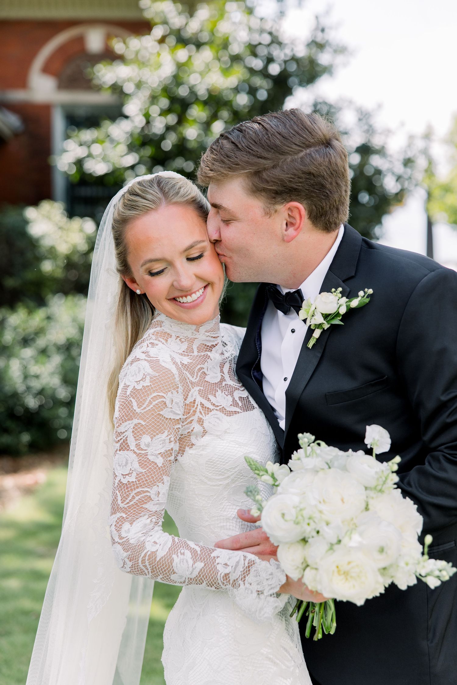 Bride in lace long-sleeve gown holds white bouquet as groom in black tuxedo kisses her cheek outdoors.