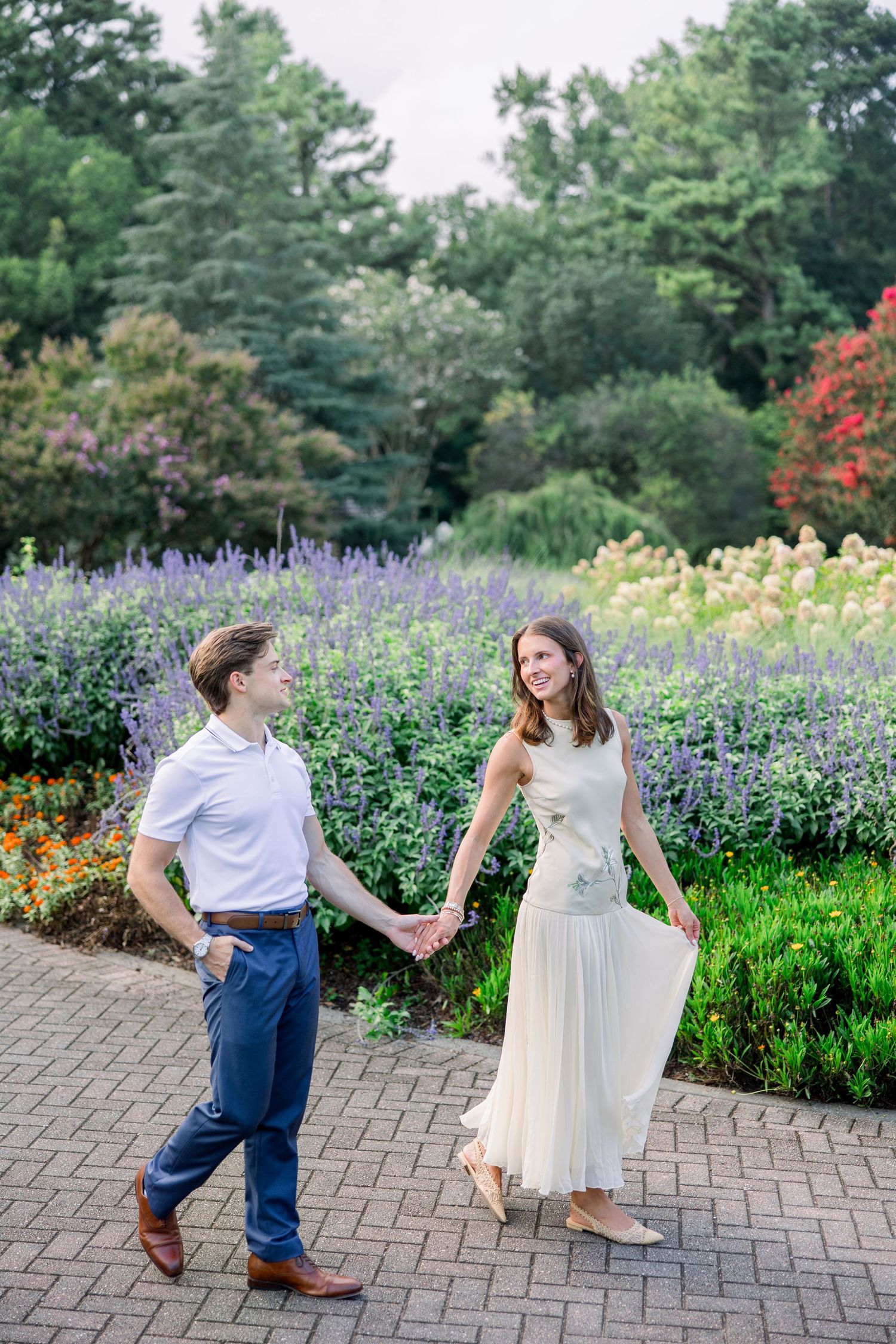 Couple holding hands and smiling while walking through a colorful garden with purple and red flowers.