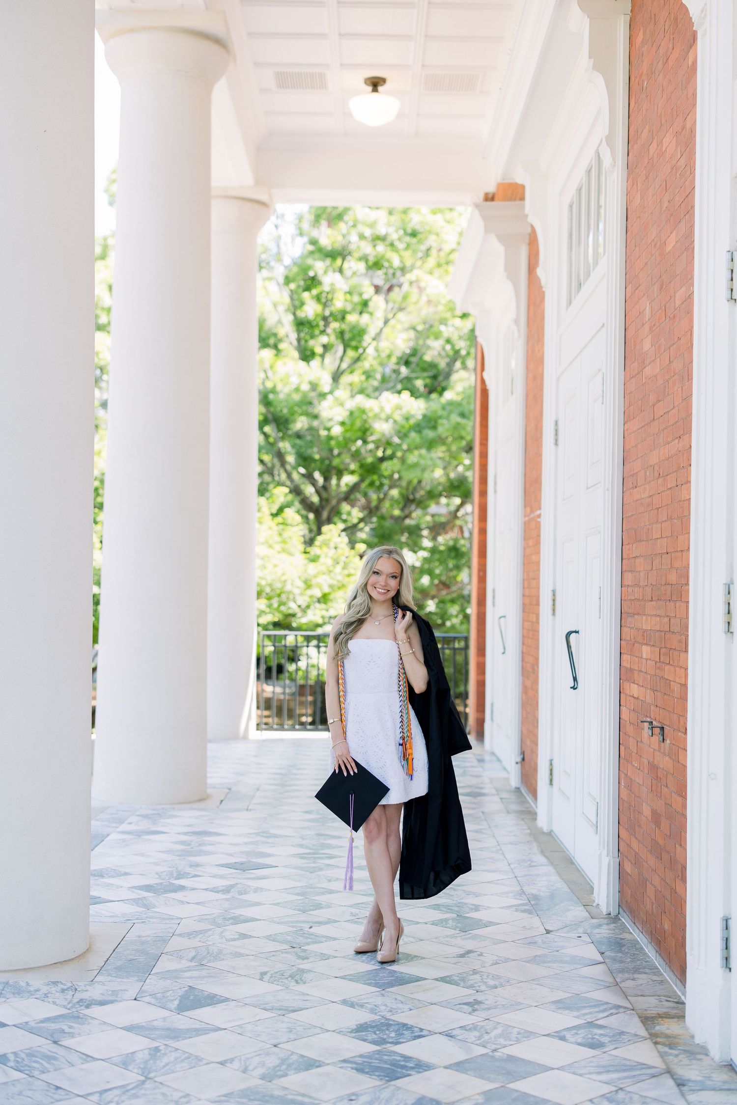 Smiling blonde woman in white dress and black graduation gown poses between white columns at a university building.