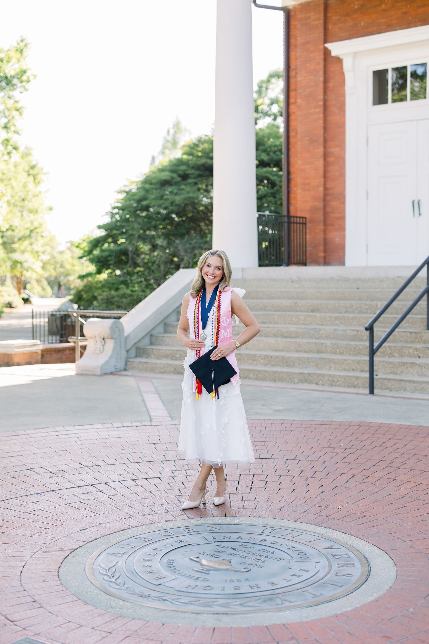 A smiling graduate in a white dress holds her cap while standing on a university seal, wearing colorful honor cords.
