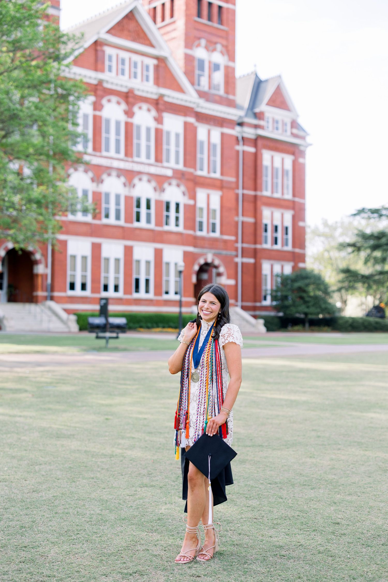 Smiling graduate in white dress with colorful honor cords holds cap outside red brick university building.