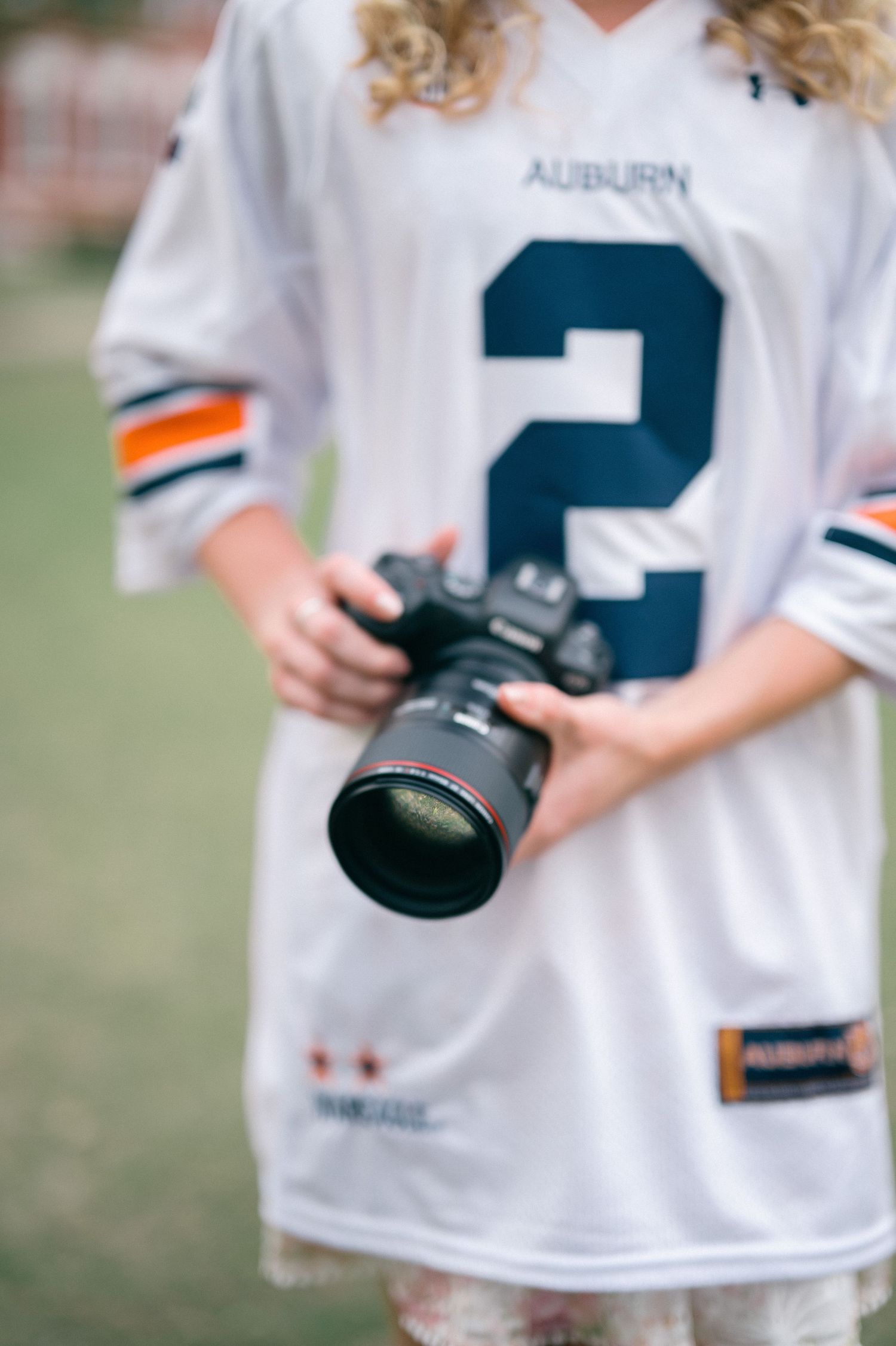 A person wearing a white jersey holds a professional camera while taking sports photographs outdoors.
