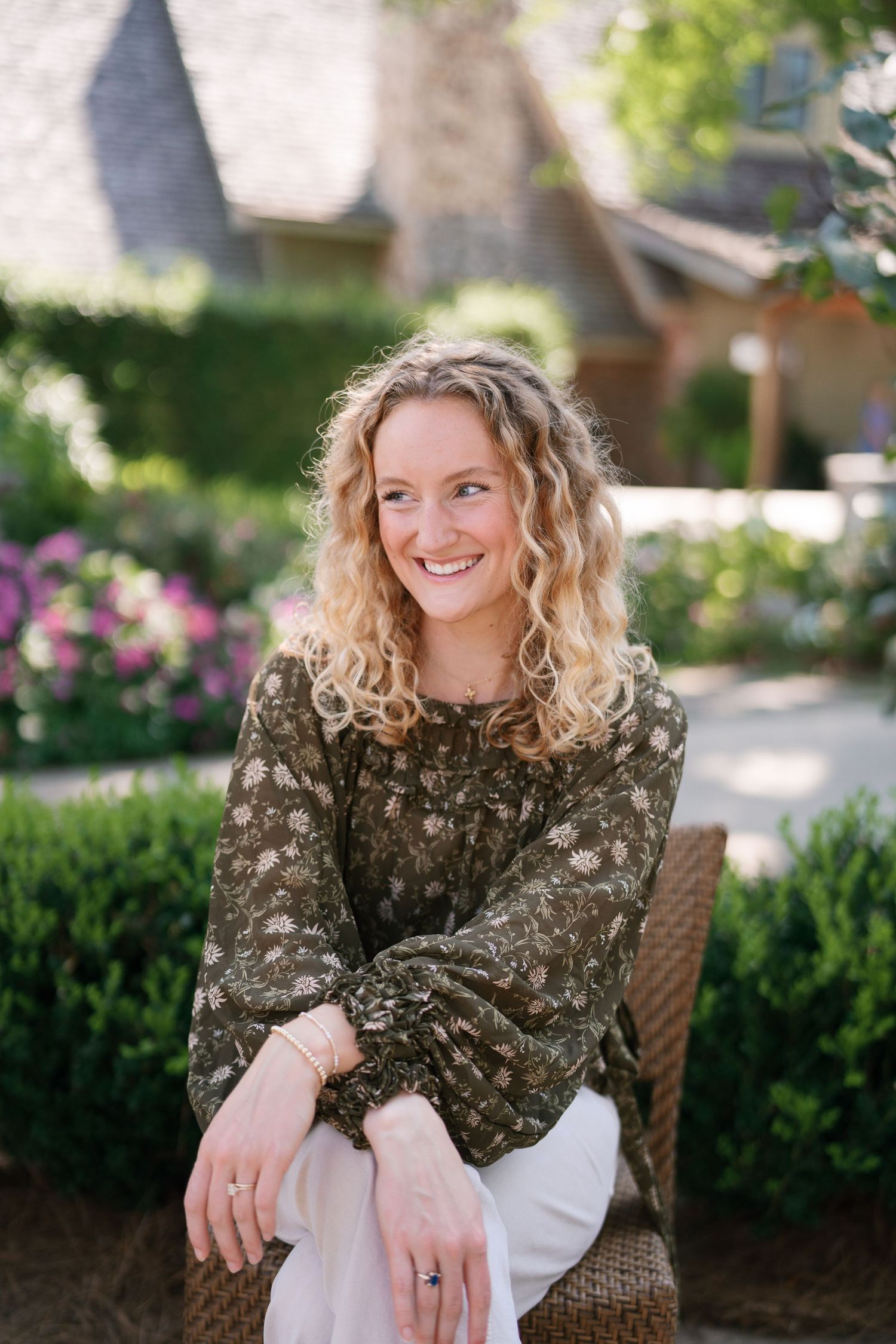 Smiling woman with curly blonde hair sits outdoors in floral green blouse and white pants, surrounded by lush garden.