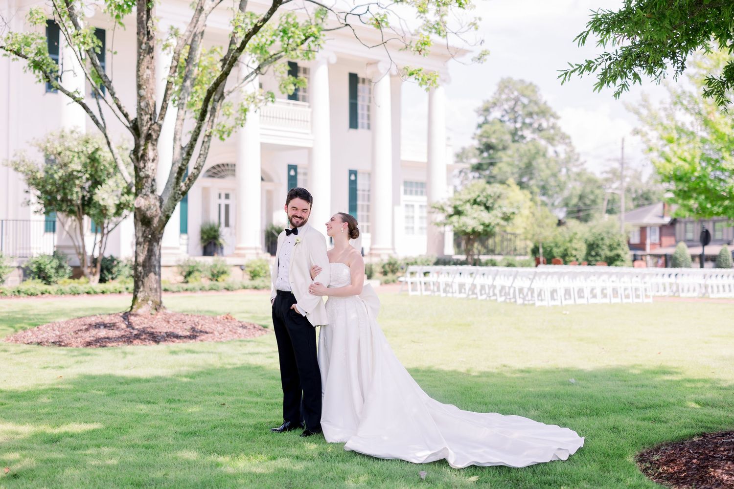 Bride and groom pose on lush green lawn before elegant white plantation-style wedding venue on sunny day.
