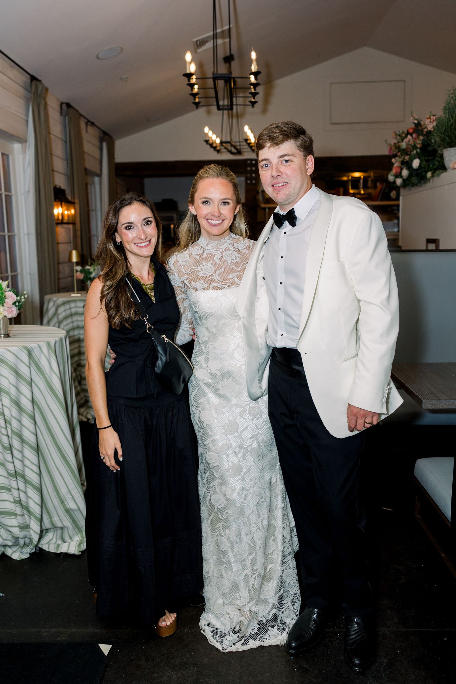 A bride in lace gown poses with guests at elegant wedding reception venue with chandelier lighting.