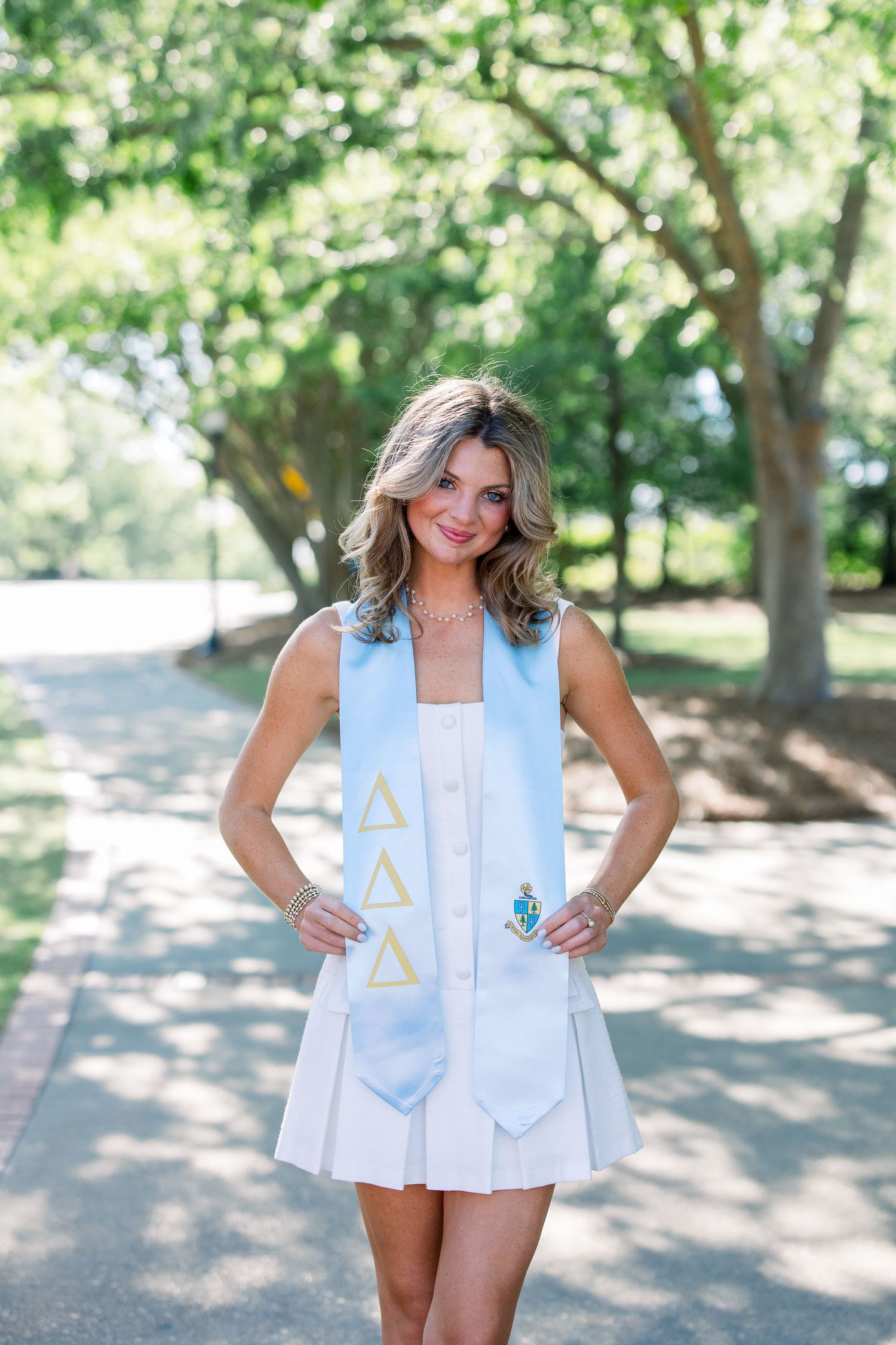 Young woman in white dress wearing Delta Delta Delta sorority graduation stole, posing outdoors on a tree-lined path.